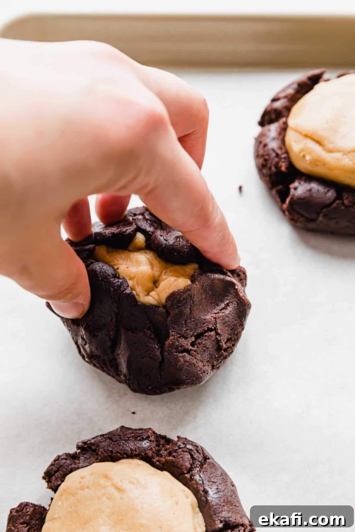 Μπισκότα με φυστικοβούτυρο και καφέ brownie Crumbl 7 Ένα χέρι τραβάει τις άκρες μιας μπάλας ζύμης μπισκότου μπράουνι πάνω από μια μπάλα ζύμης φυστικοβούτυρου.