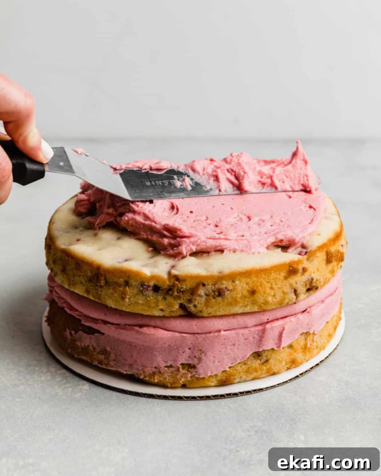 Perfect Lemon Raspberry Cake 10 A spatula spreading a pink raspberry frosting overtop two layer cake.