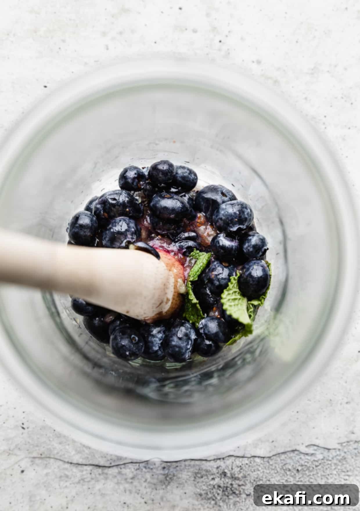 Blueberry Bliss Quencher 5 A wood muddler pressing down on fresh blueberries and mint leaves in a glass, demonstrating the muddling process for a mocktail.