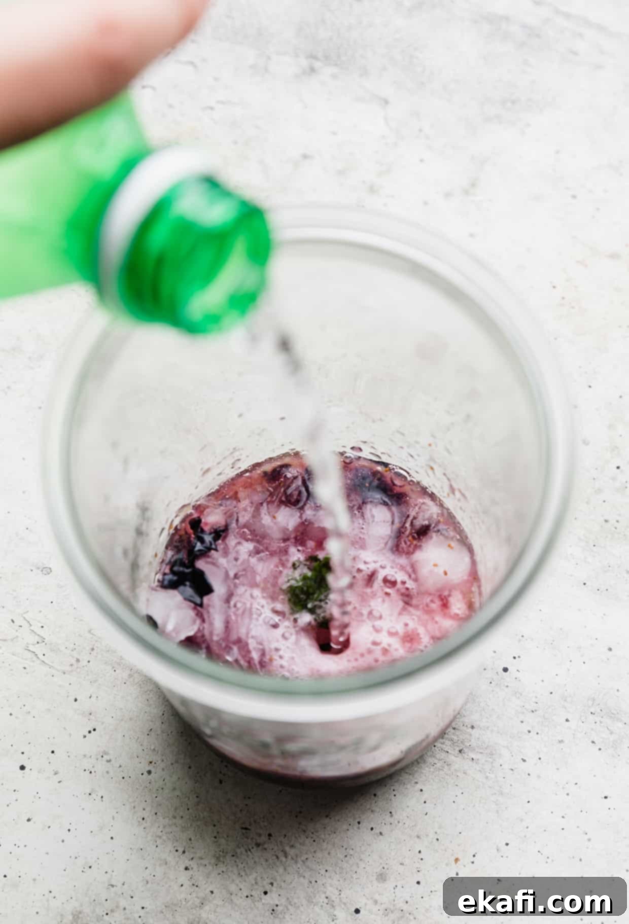 Blueberry Bliss Quencher 7 Clear soda being poured into a glass jar with mashed blueberries and mint, forming a virgin blueberry mojito.