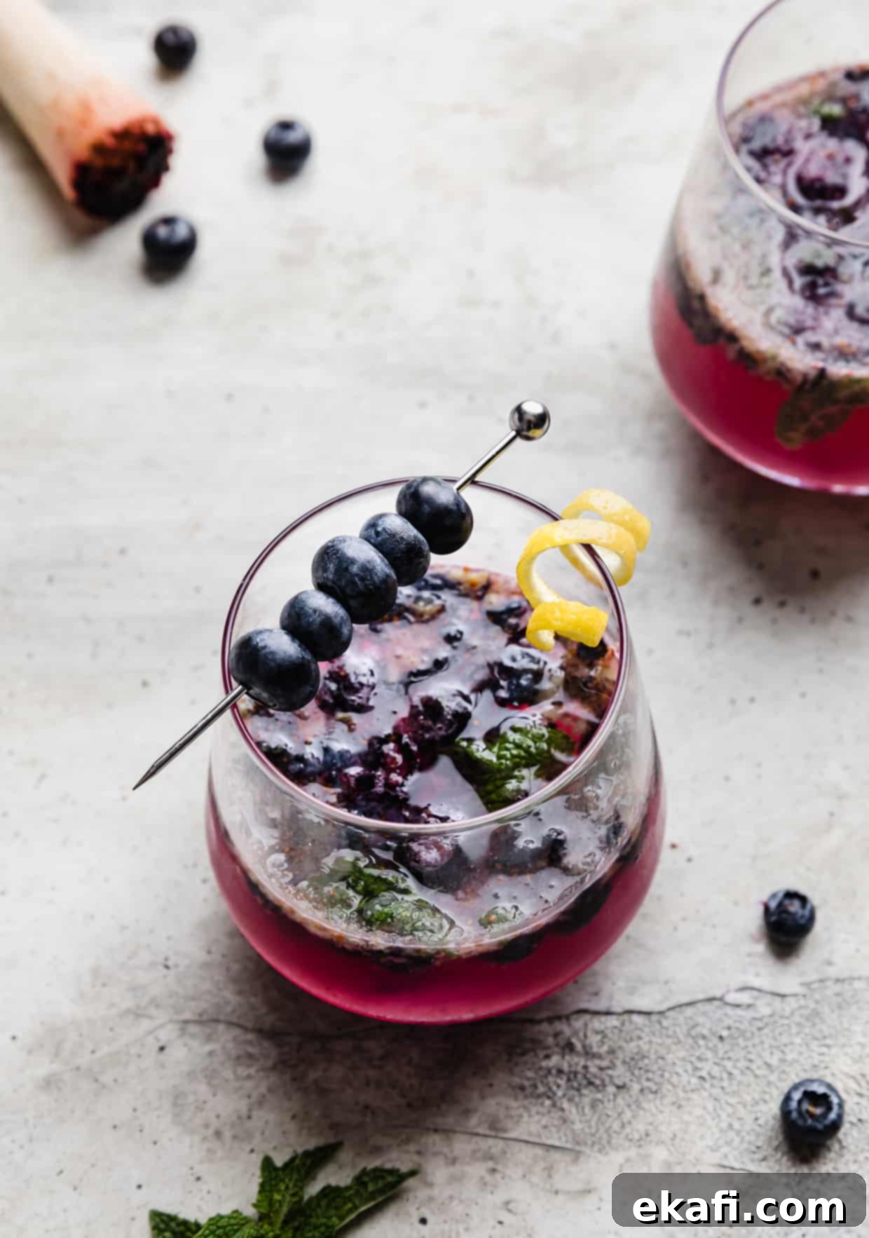 Blueberry Bliss Quencher 8 A tall glass cup filled with a vibrant blueberry mocktail, garnished with fresh blueberries and mint, set against a bright background.