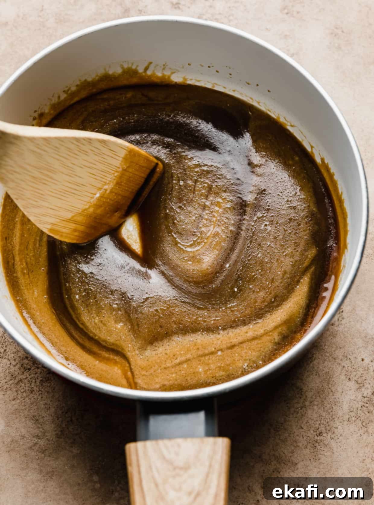 Salted Butterscotch Sauce 6 Salted Butterscotch Sauce in a white pan with a wooden spoon stirring the mixture, demonstrating its rich, smooth texture.