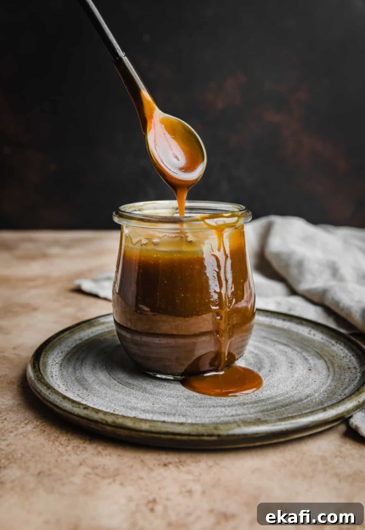 Salted Butterscotch Sauce 7 A spoon lifting out a generous portion of Salted Butterscotch topping from a small glass jar, against a dark brown background, highlighting its rich color and texture.