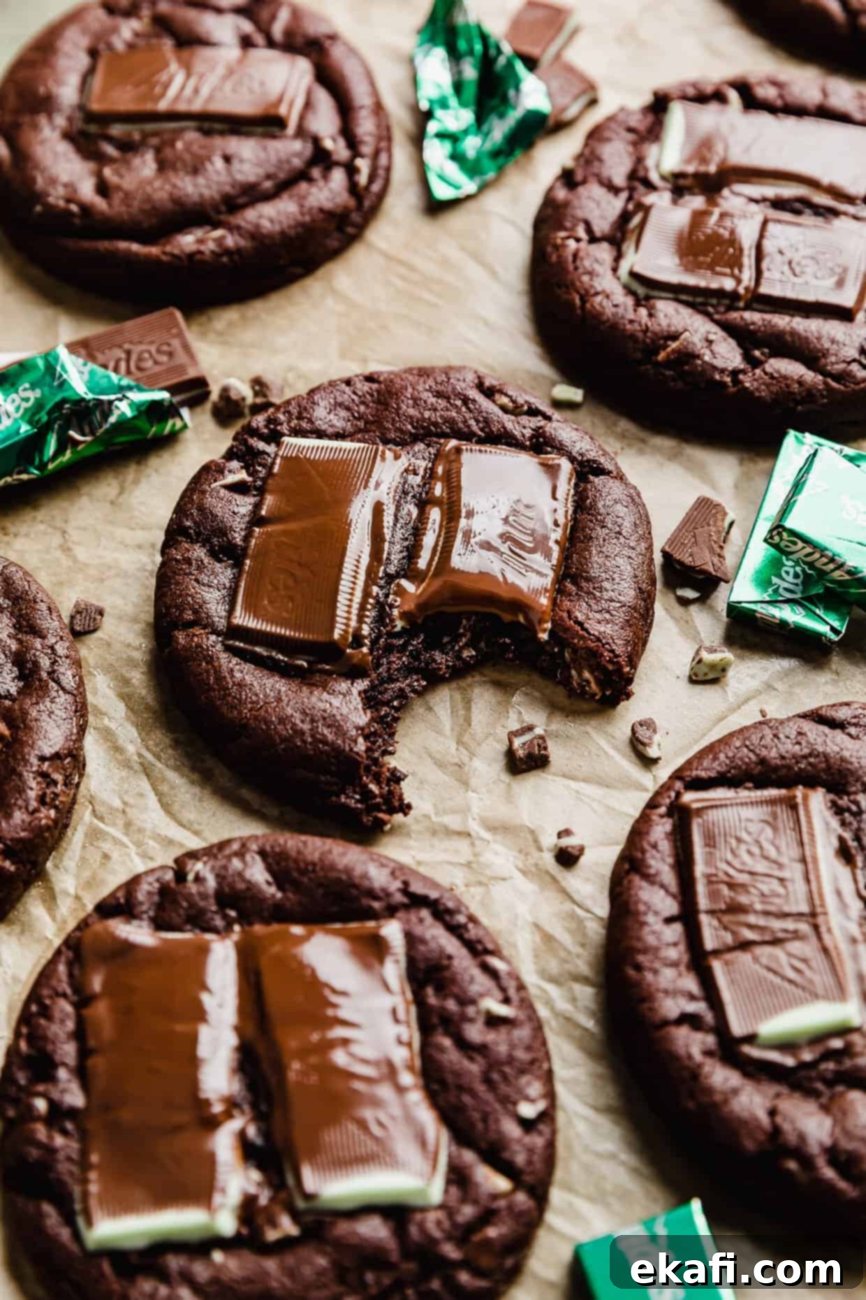 Decadent Andes Mint Chocolate Cookies 7 A bite taken out of a Chocolate Andes Mint Cookie on a tan background with Andes mint candies surrounding the cookie.