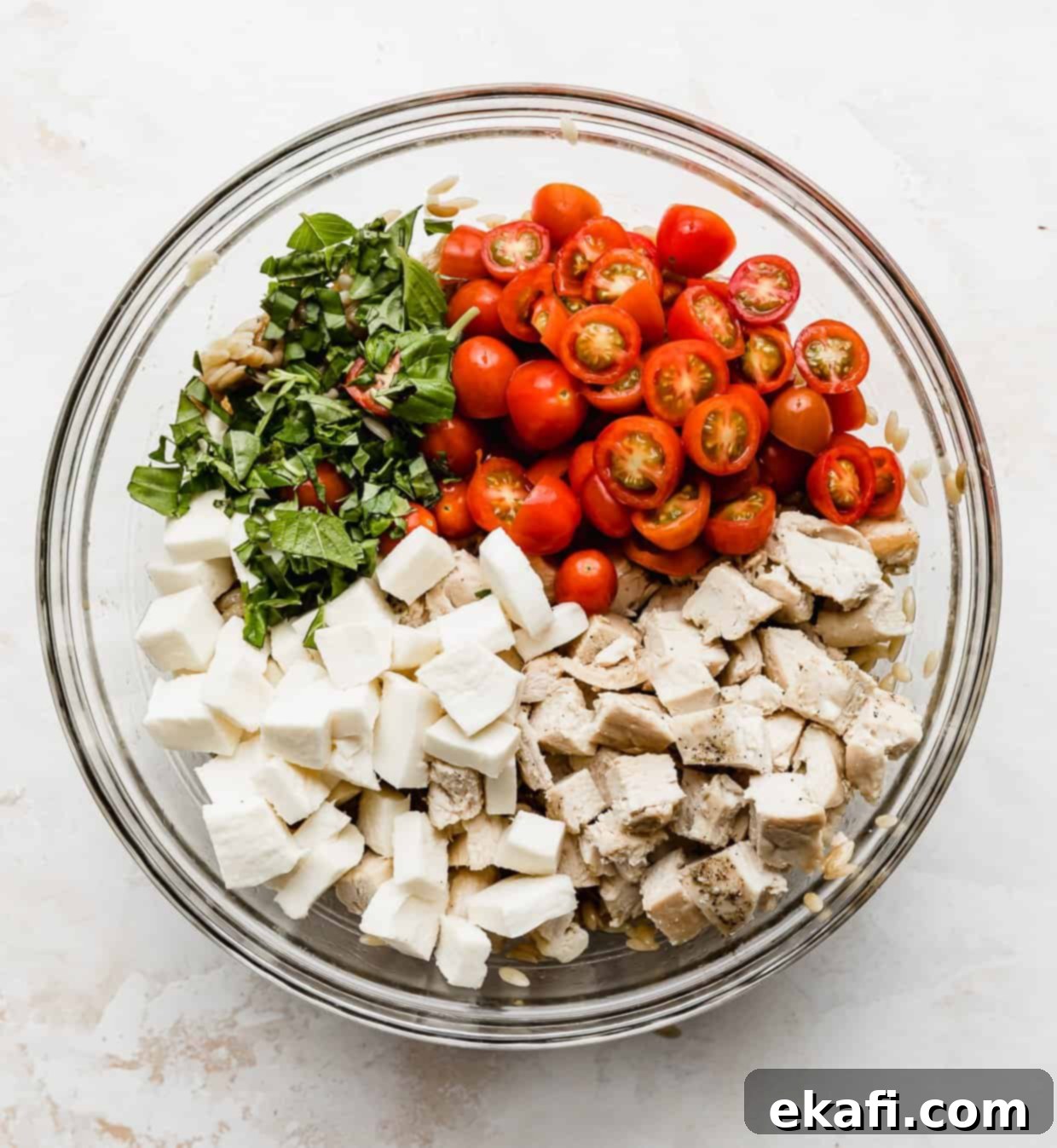 Perfect Summer Caprese Orzo 7 Glass bowl with orzo caprese salad ingredients in it: mozzarella, tomatoes, basil, chopped chicken, orzo pasta.