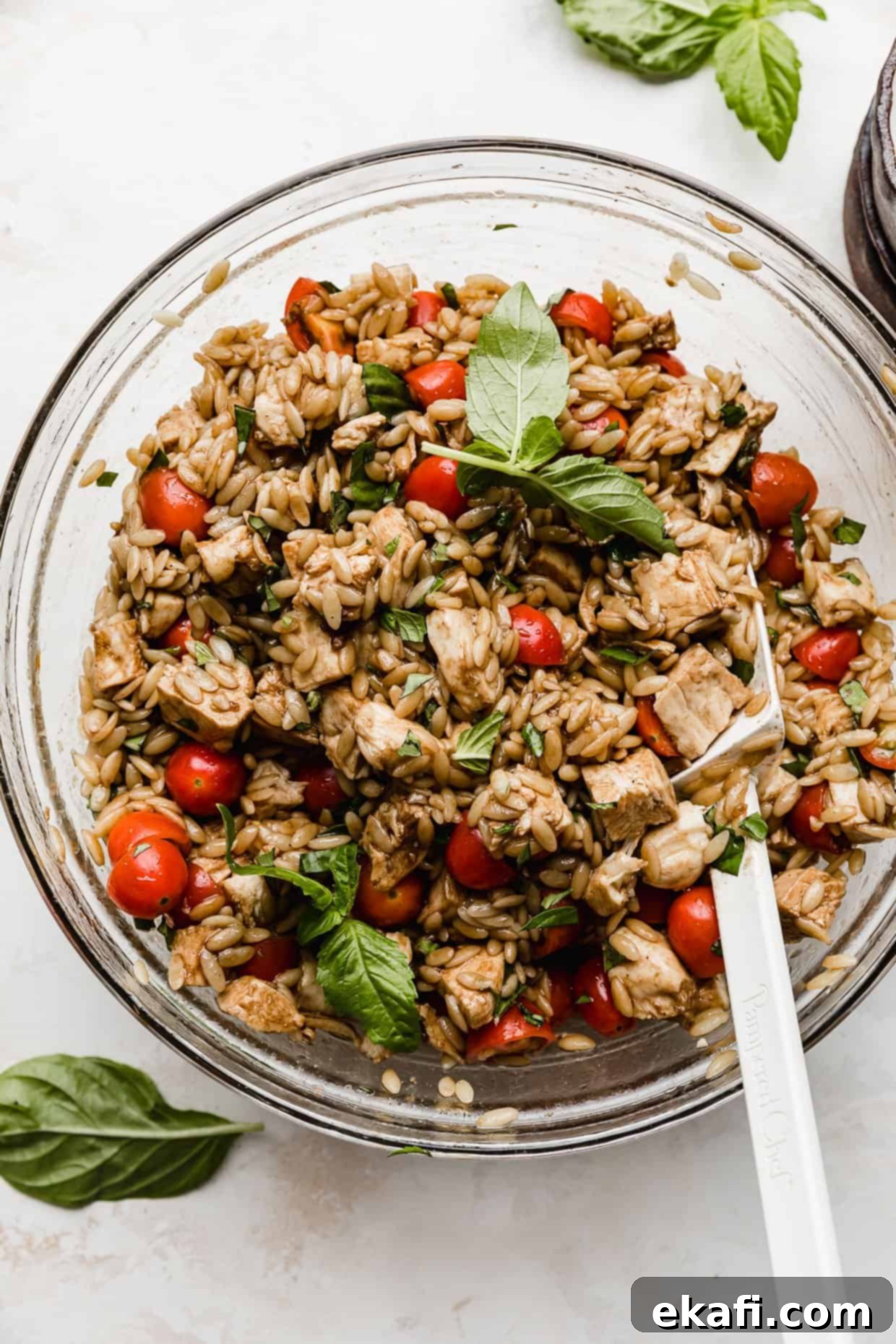 Perfect Summer Caprese Orzo 8 Chicken Caprese Orzo Salad in a glass bowl with a white spatula stirring the salad.