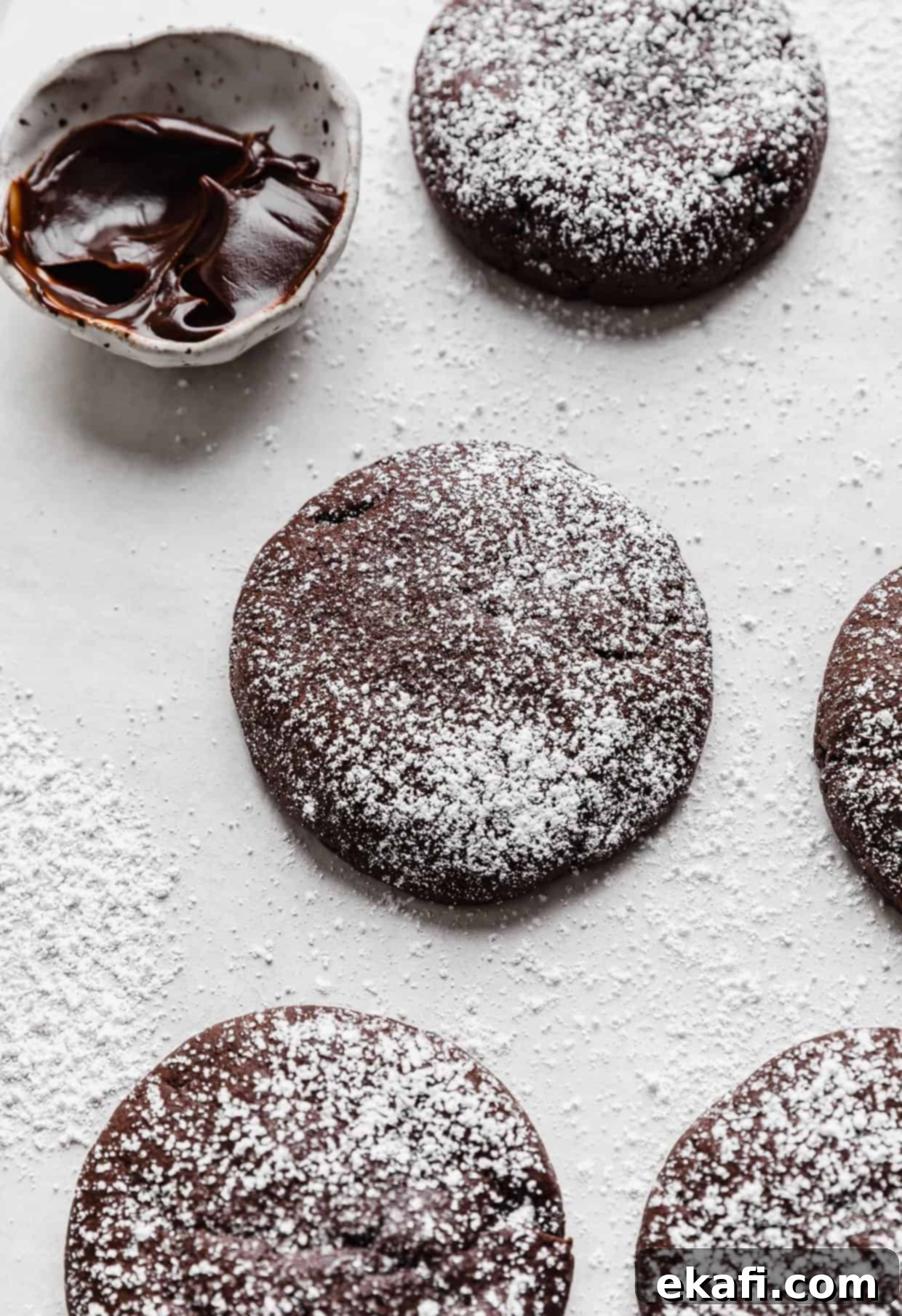 Μπισκότα με Καρδιά Λάβας 11 Crumbl Molten Lava Cookies πασπαλισμένα με ζάχαρη άχνη σε λευκό φόντο με ένα μικρό μπολ ζεστής φοντάν στο παρασκήνιο.