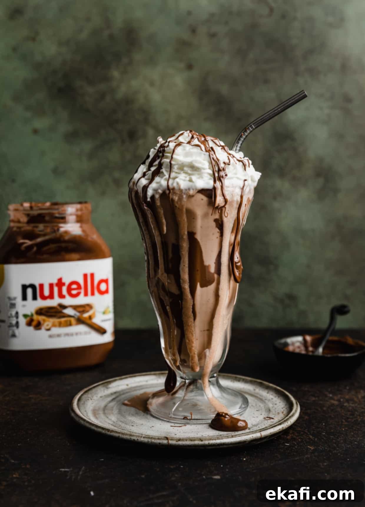 Μιλκσέικ Νουτέλας 2 Ένα γυάλινο ποτήρι milkshake με Nutella Milkshake, γαρνιρισμένο με σαντιγί και σιρόπι Nutella, μπροστά από ένα σκούρο πράσινο φόντο.