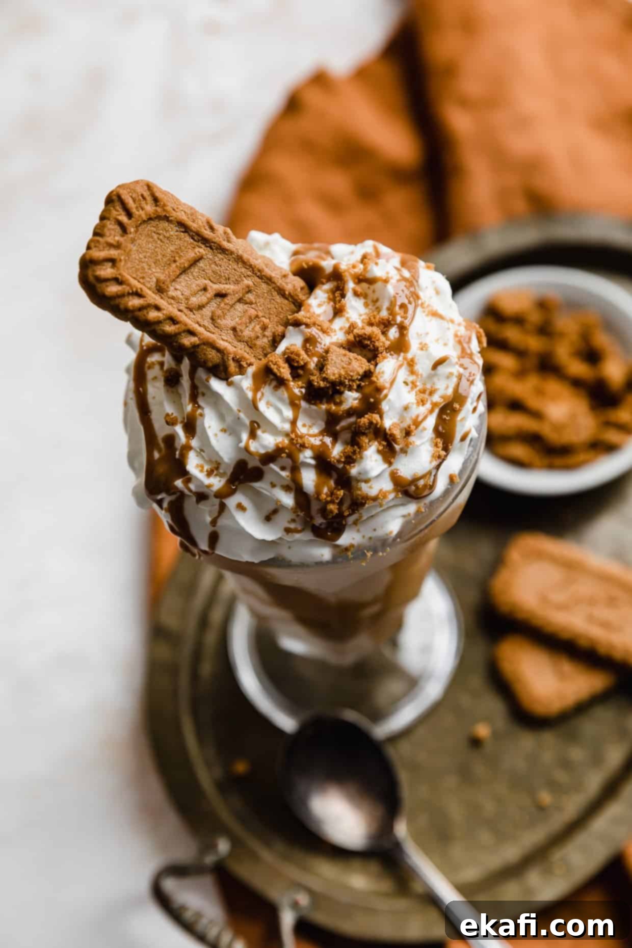 Biscoff Milkshake 2 A rich Biscoff Milkshake topped with a generous swirl of fluffy whipped cream, a delicate Biscoff drizzle, and a whole Lotus Biscoff cookie. The inviting golden-brown drink is presented in a tall glass on a stylish bronze tray, set against a pristine white background, perfect for a luxurious dessert.