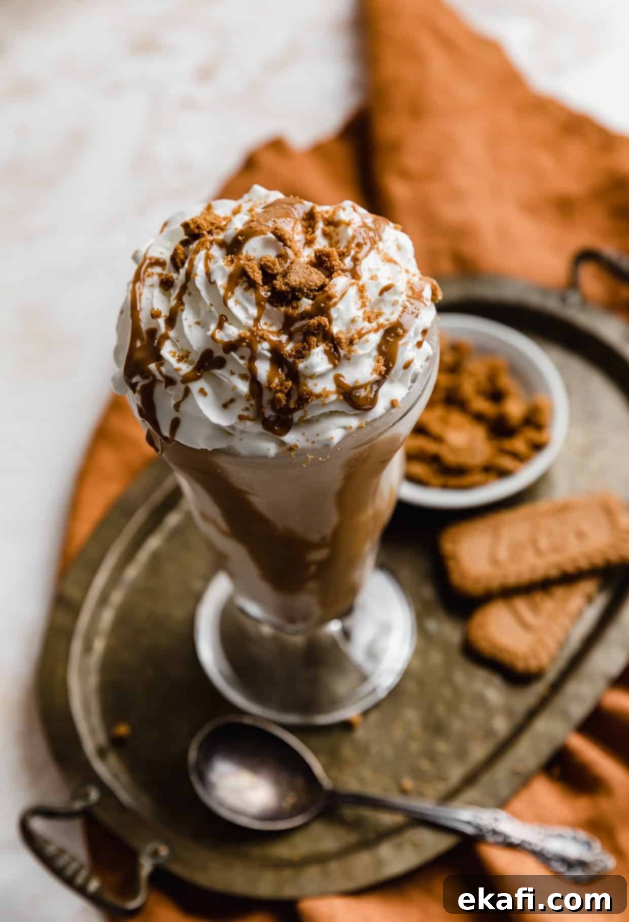 Biscoff Milkshake 6 A perfectly prepared Biscoff Milkshake in a clear glass cup, adorned with whipped cream and a sprinkle of crushed Biscoff cookies, presented on an elegant bronze tray. In the soft-focus background, whole Biscoff cookies are artfully arranged, completing the indulgent and inviting scene.