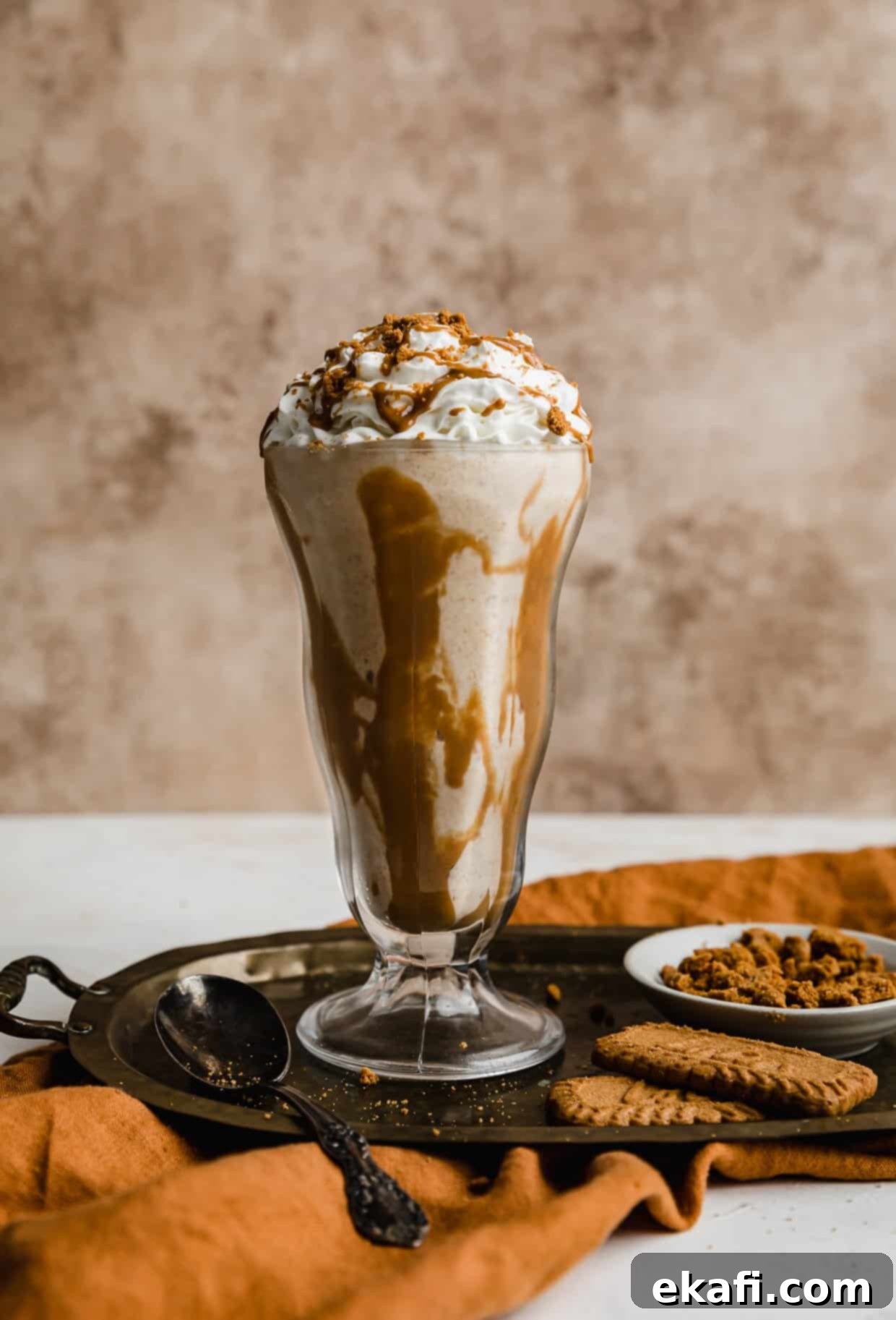 Biscoff Milkshake 7 A beautifully presented Biscoff Milkshake in a stylish glass cup, garnished with a generous dollop of whipped cream and a sprinkle of crushed Biscoff cookies. The delightful drink is set against a warm, light brown textured background, with soft lighting highlighting its inviting creaminess.