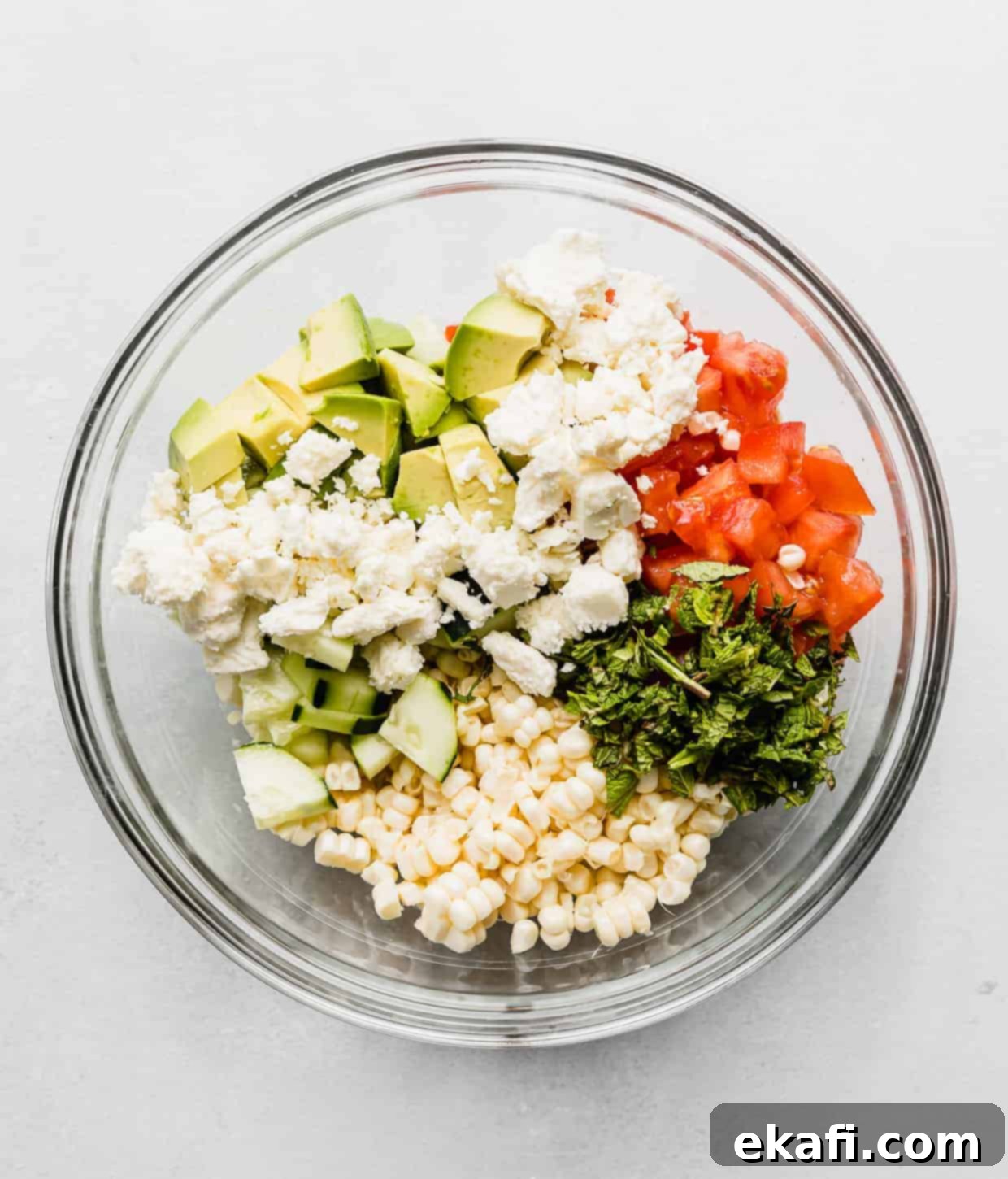 Creamy Zesty Corn Avocado Feta Salad 4 A clear glass bowl filled with colorful ingredients: raw corn kernels, diced red tomatoes, crumbled white feta cheese, cubed green avocado, and fresh mint leaves, awaiting dressing.