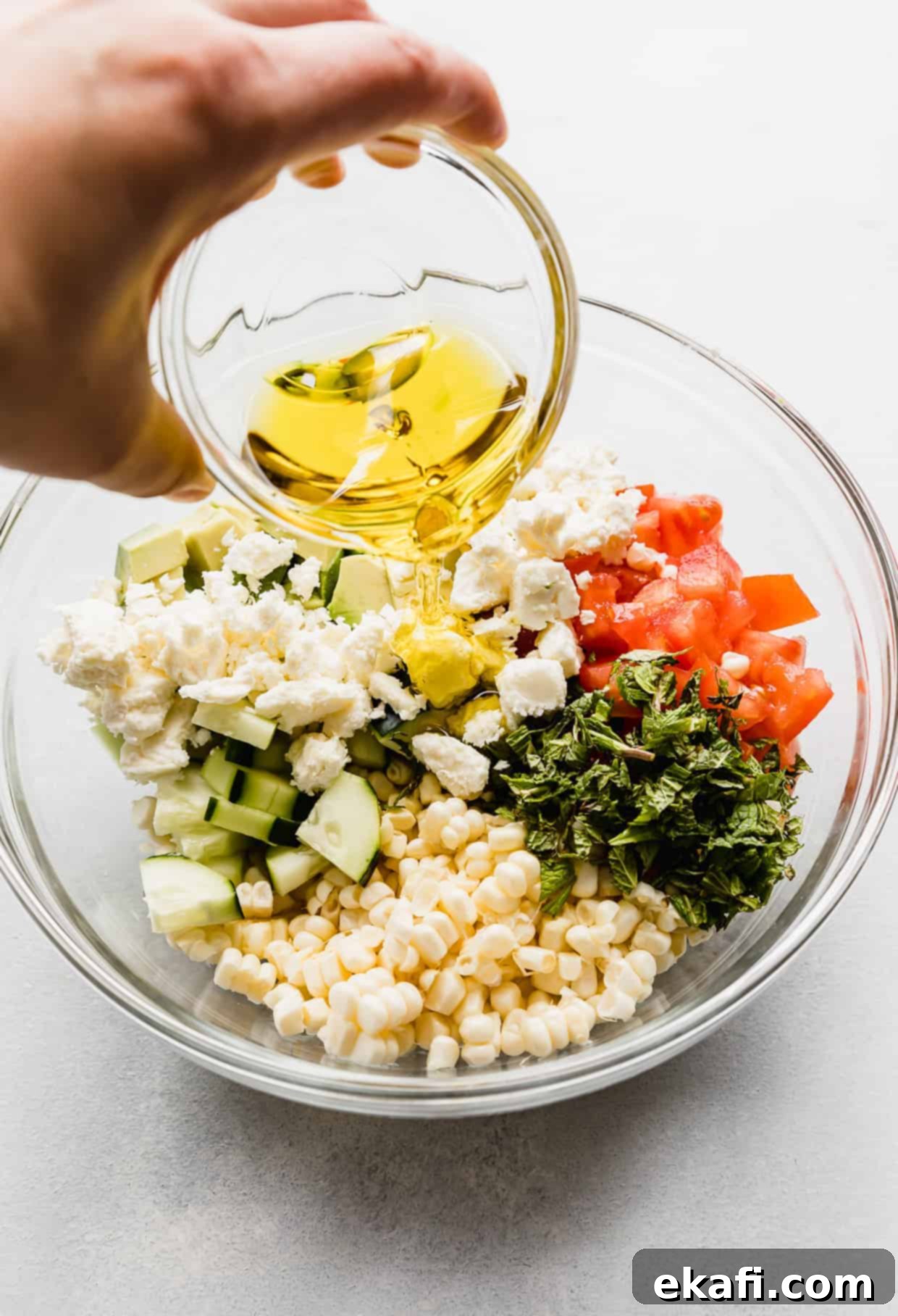 Creamy Zesty Corn Avocado Feta Salad 5 A stream of golden olive oil being poured over the vibrant mix of feta corn salad ingredients in a glass bowl.
