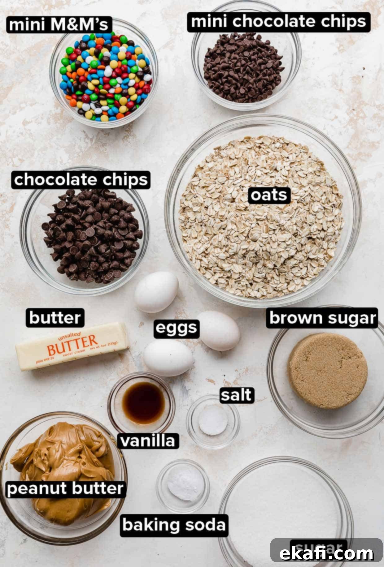 Loaded Chewy Cookies 3 Individual ingredients for monster cookies, including butter, sugars, eggs, peanut butter, oats, chocolate chips, and M&M's, portioned into glass bowls on a clean white surface.