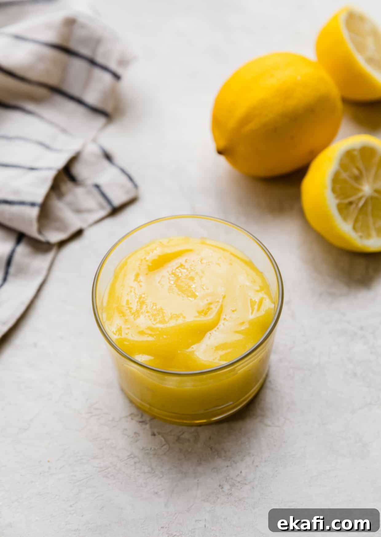 Luscious Lemon Curd Perfection 2 A homemade Lemon Curd Recipe in a glass cup on a light gray background with a full lemon and two lemon halves in the background.