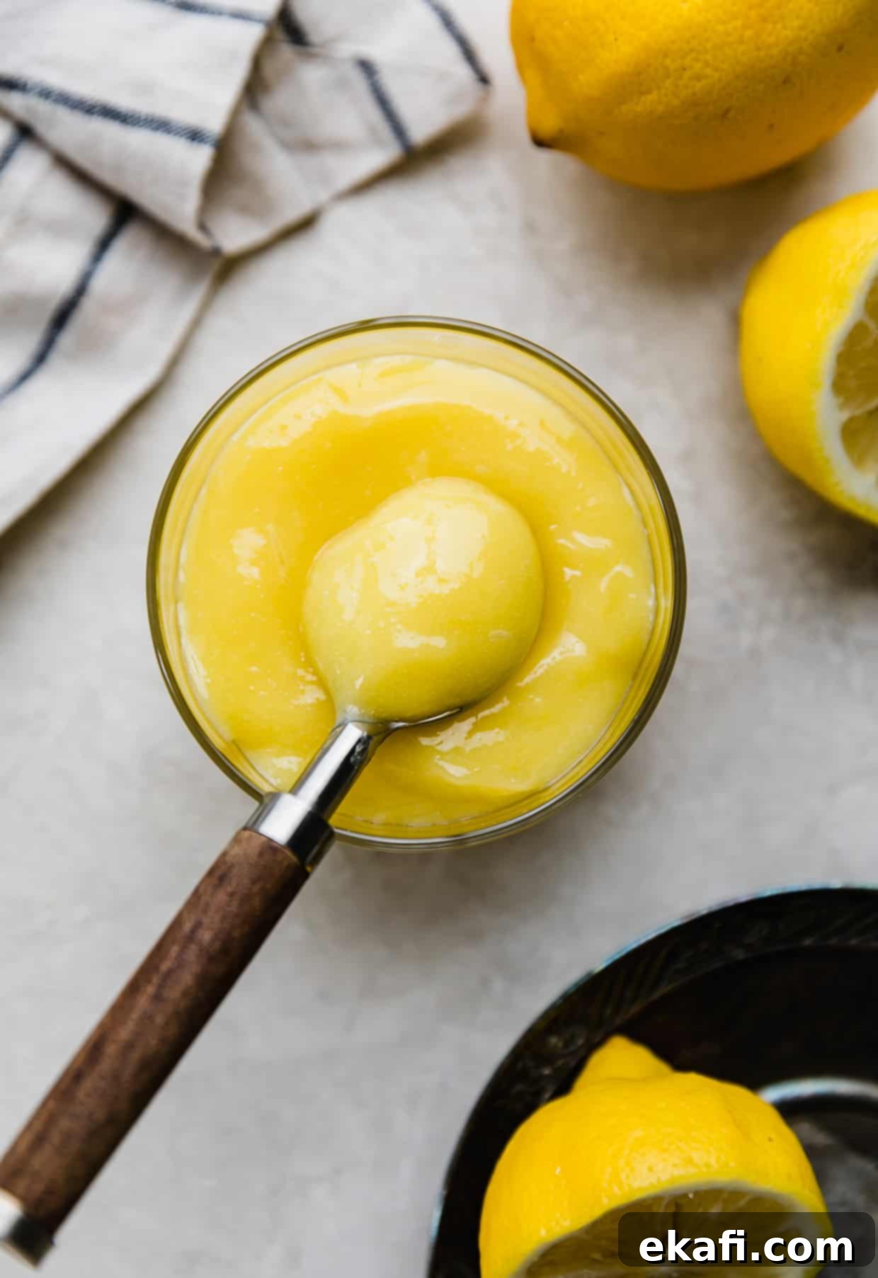 Luscious Lemon Curd Perfection 7 Overhead photo of yellow lemon curd in a glass cup on a light gray background, with a brown spoon scooping up a portion of the lemon curd.