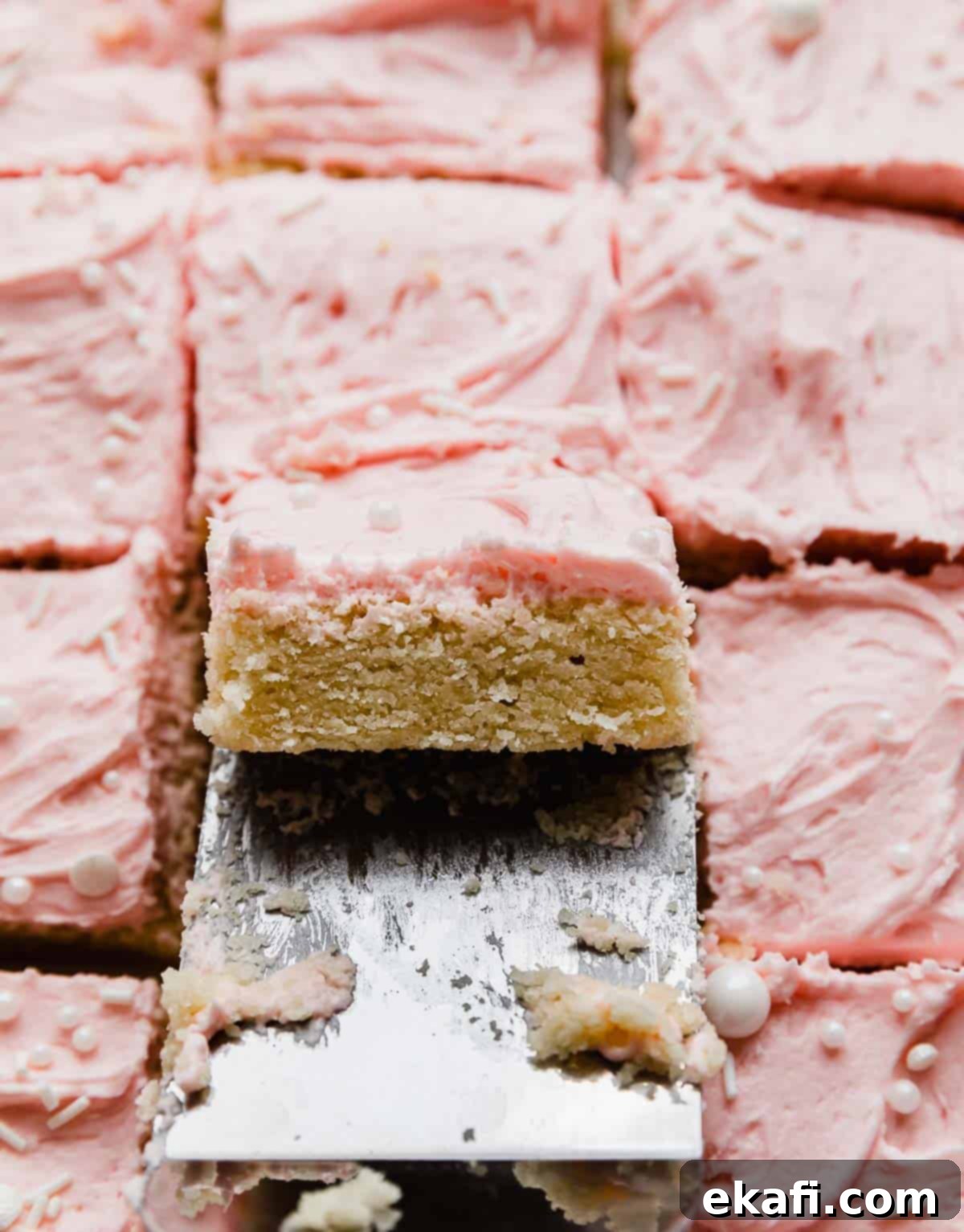 Frosted Sugar Cookie Bars 7 A metal spatula lifting up a square of Sugar Cookie Bar that's frosted with pink buttercream frosting, showcasing its delightful softness.