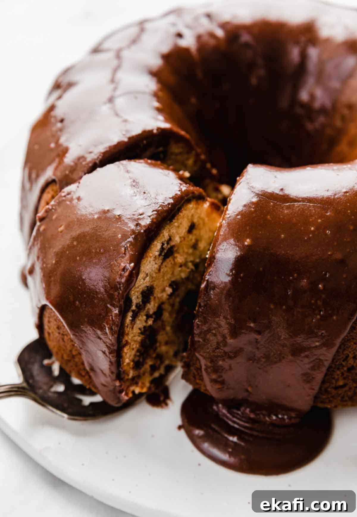 Rich Chocolate Chip Bundt Perfection 3 Chocolate frosting covered Chocolate Chip Bundt Cake on a white plate with a slice being removed from the cake, highlighting the inviting interior.