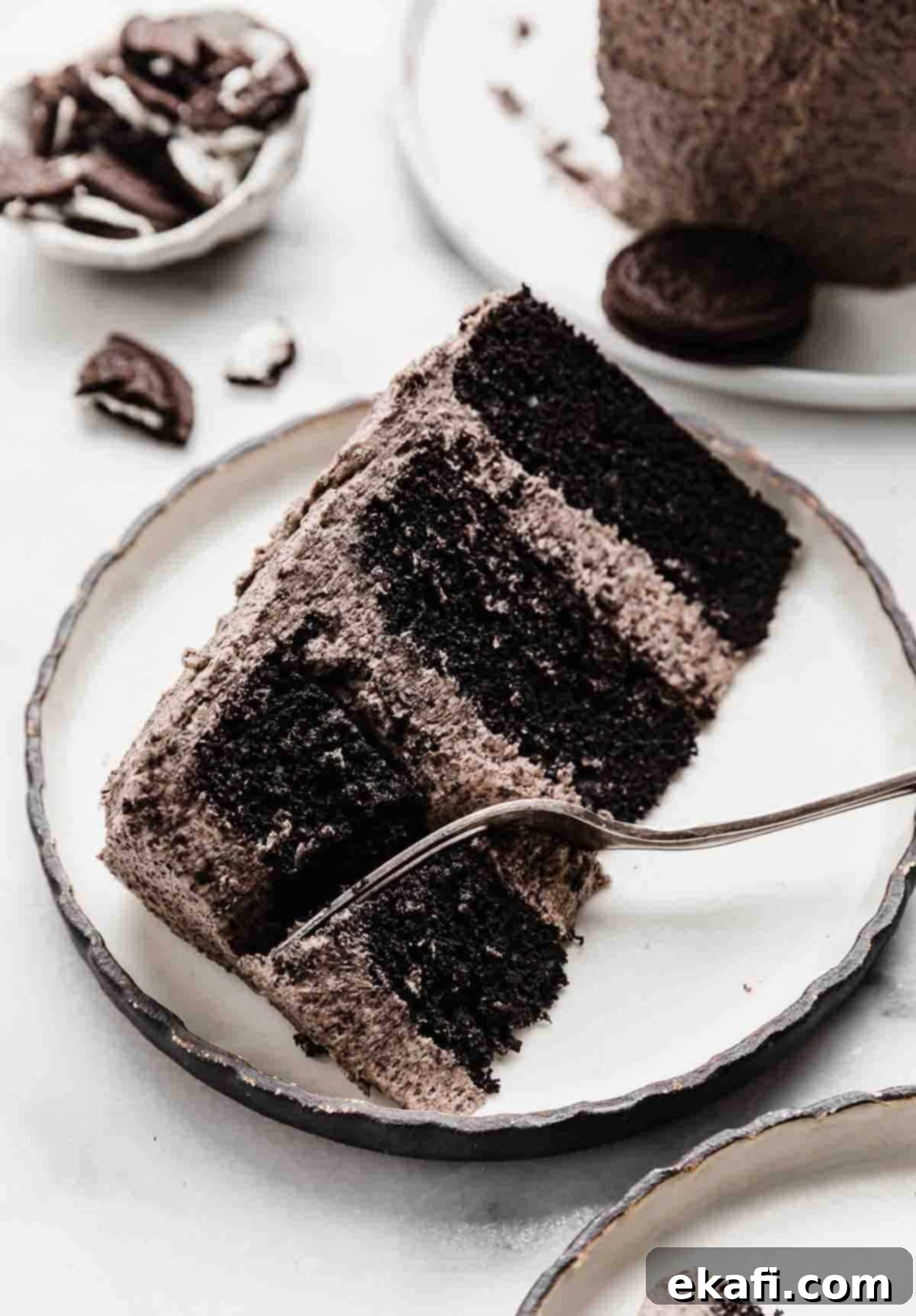 Decadent Oreo Dream 7 A slice of three tiered oreo cake on a black rimmed white plate with a fork cutting into the top portion of the cake.