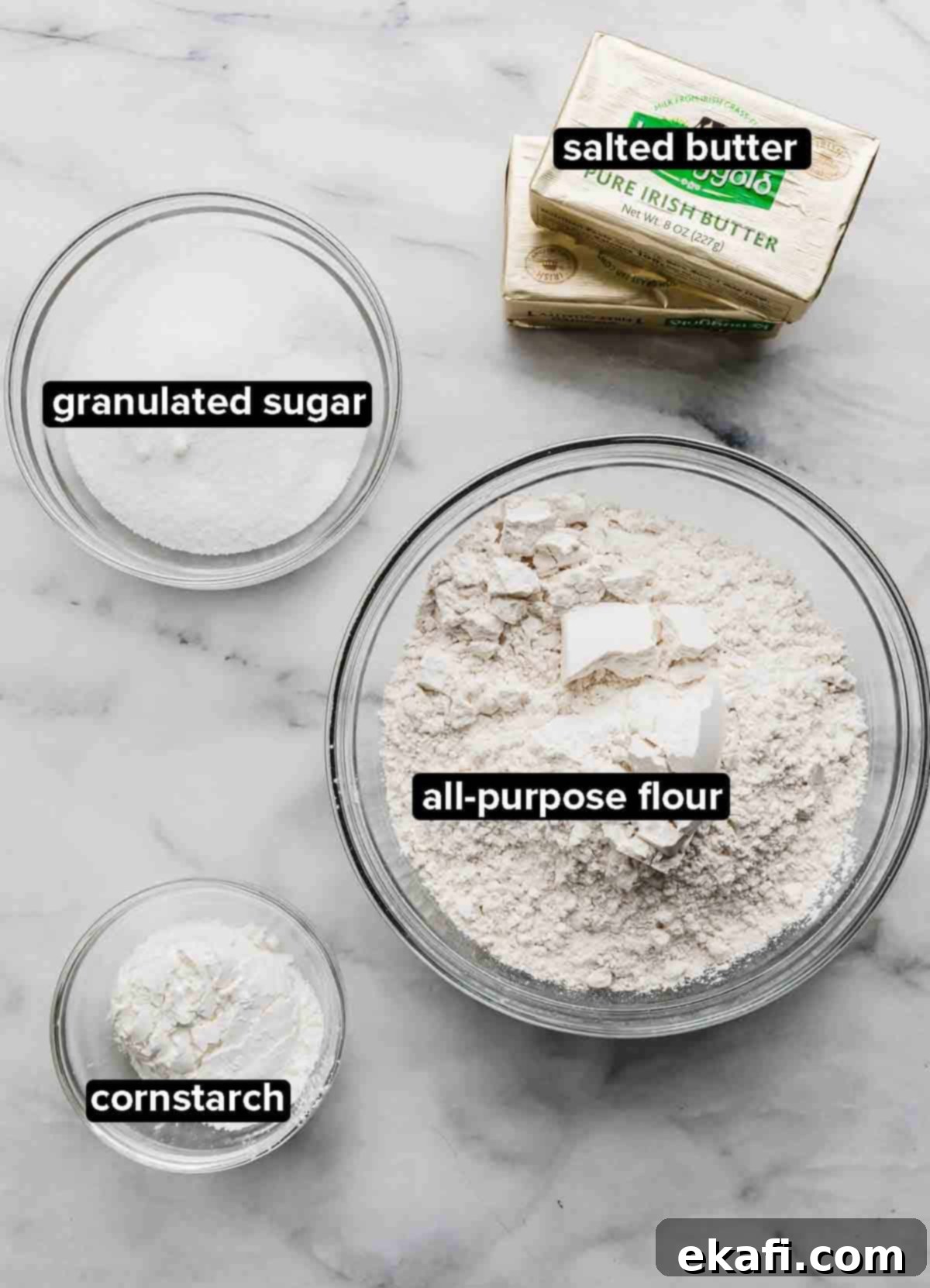 Grandma's Buttery Shortbread 3 Grandma's Shortbread Cookie ingredients portioned into glass bowls on a white and gray marble background: flour, sugar, cornstarch, and butter.