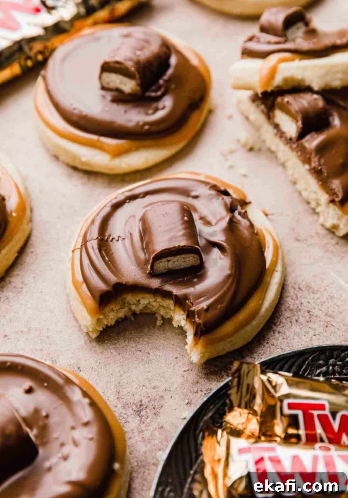 Τουίξ Μπισκότα 2 Μπισκότα Twix φτιαγμένα με βάση μπισκότου βουτύρου, επικαλυμμένα με καραμέλα, σοκολάτα και μια μπάρα Twix, σε ανοιχτό καφέ φόντο.