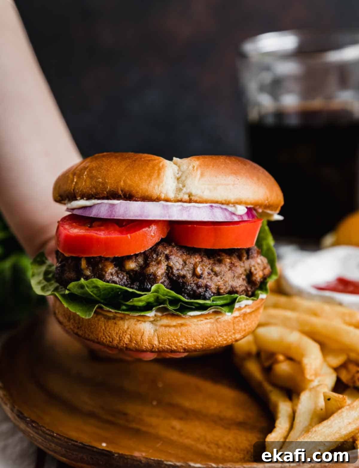 Μπέργκερ Μπριζόλας 2 Ένα χέρι κρατάει ένα Steakhouse Burger σε ψωμάκι με μαρούλι, ντομάτα και μοβ κρεμμύδι, σε σκούρο καφέ φόντο με τηγανητές πατάτες στο παρασκήνιο.