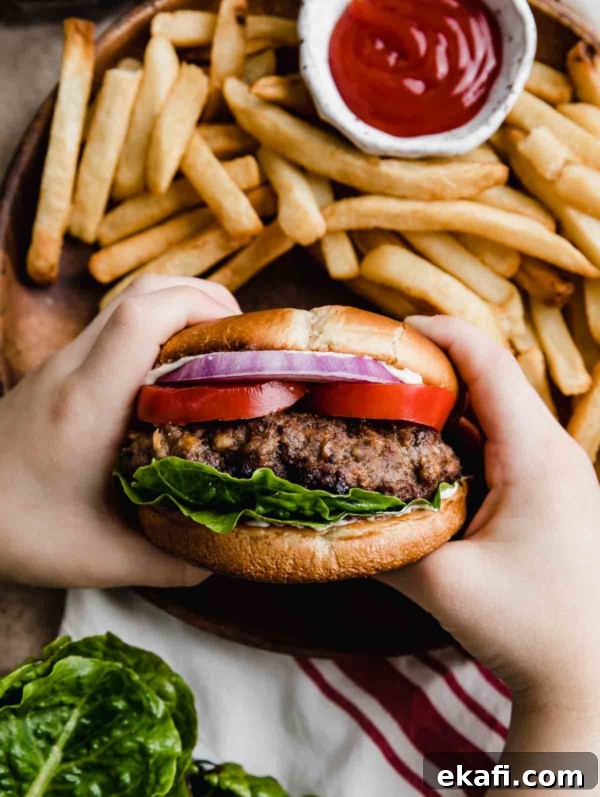 Μπέργκερ Μπριζόλας 6 Δύο χέρια κρατούν ένα Steakhouse Burger σε ψωμάκι με μαρούλι, ντομάτα και μοβ κρεμμύδι, με τηγανητές πατάτες στο παρασκήνιο.