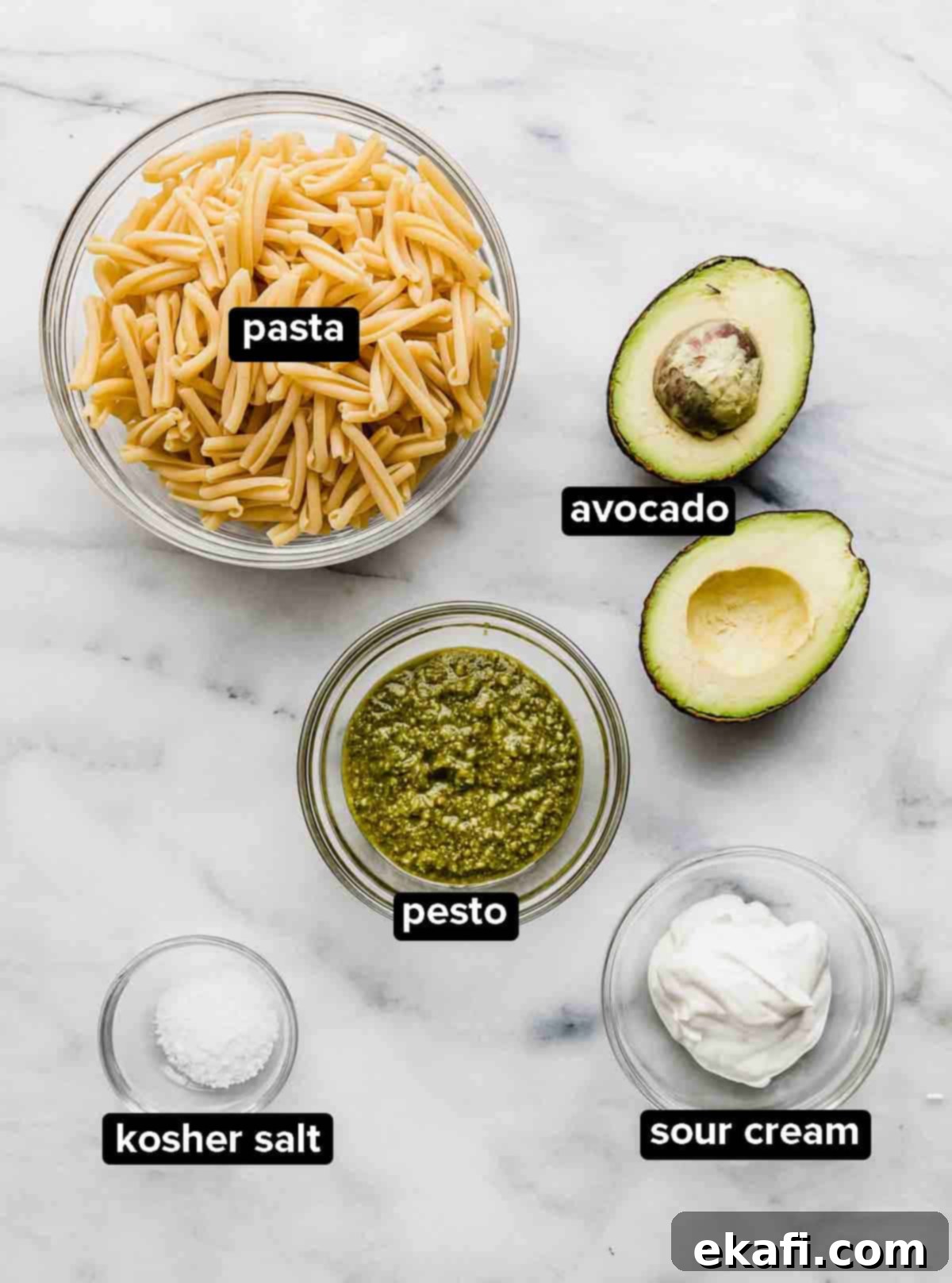 Vibrant Avocado Pesto Pasta 3 Avocado Pesto Pasta ingredients on a white and gray marble background: noodles, sour cream, salt, basil pesto, and an avocado.