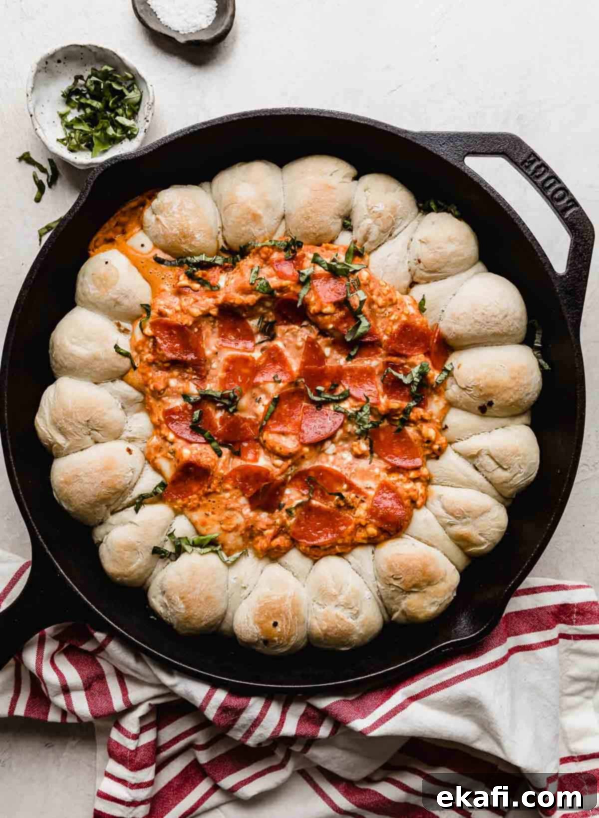 This Pepperoni Pizza Dip is cheesy, creamy, and bursting with all the pizza flavors! It's an easy appetizer recipe loved by all! Pepperoni Pizza Dip in a black cast iron skillet on a rustic white background, garnished with fresh basil. The dip is bubbling, and the pull-apart dough rolls are golden brown.