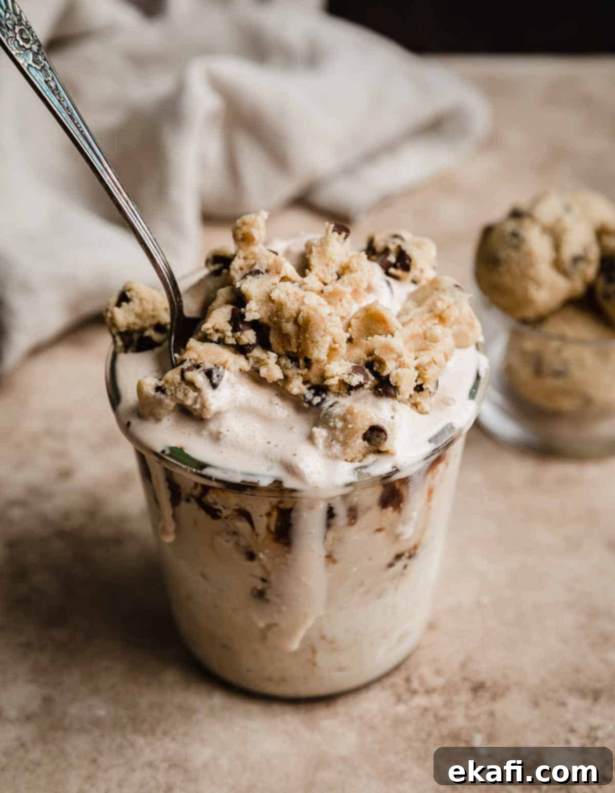 Δίνη Μπισκοτοζύμης Σοκολάτας 5 Chocolate Chip Cookie Dough Blizzard γαρνιρισμένο με κομμάτια ζύμης μπισκότων σε γυάλινο ποτήρι, σε ανοιχτό καφέ φόντο.