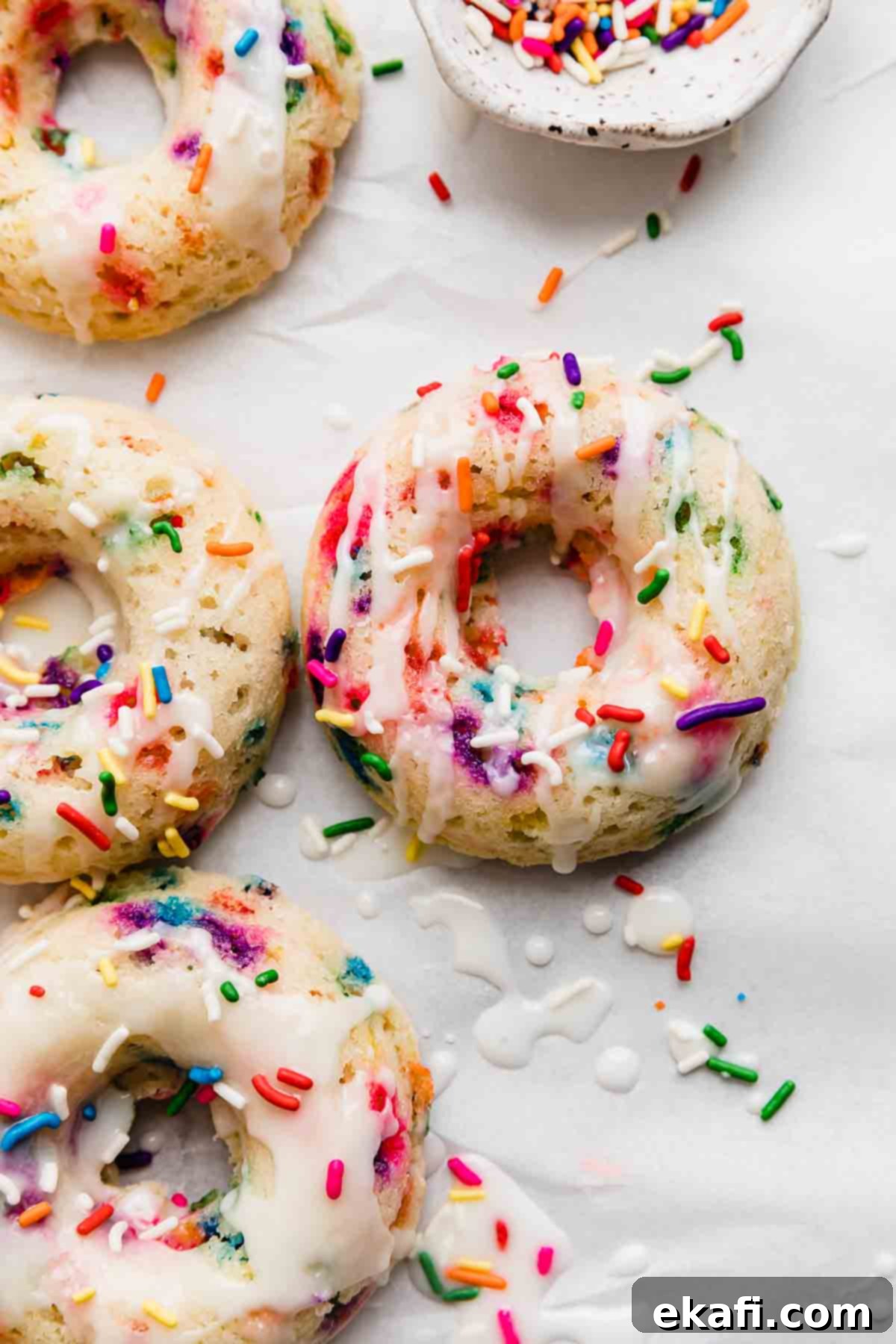 Confetti Delights 2 Four Sprinkle Donuts topped with a glaze and colorful sprinkles on a white background.