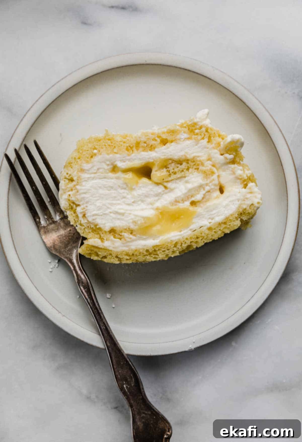 Ρολό λεμονιού 7 Μια φέτα Lemon Swiss Roll γεμάτη με κρέμα και λεμονόκρεμα, σε ένα λευκό πιάτο με ένα πιρούνι αριστερά από το κέικ.