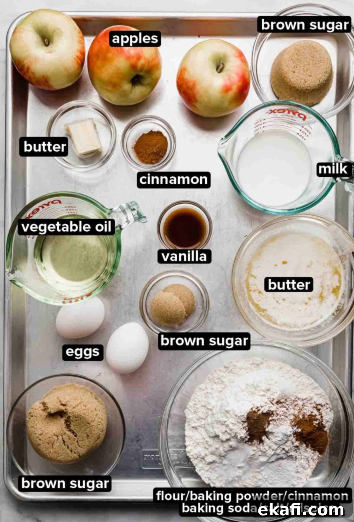 Baked Apple Fritter Delight 3 Apple Fritter Cake ingredients portioned into glass bowls on a silver baking sheet: apples, brown sugar, flour, eggs, oil, vanilla, and spices.