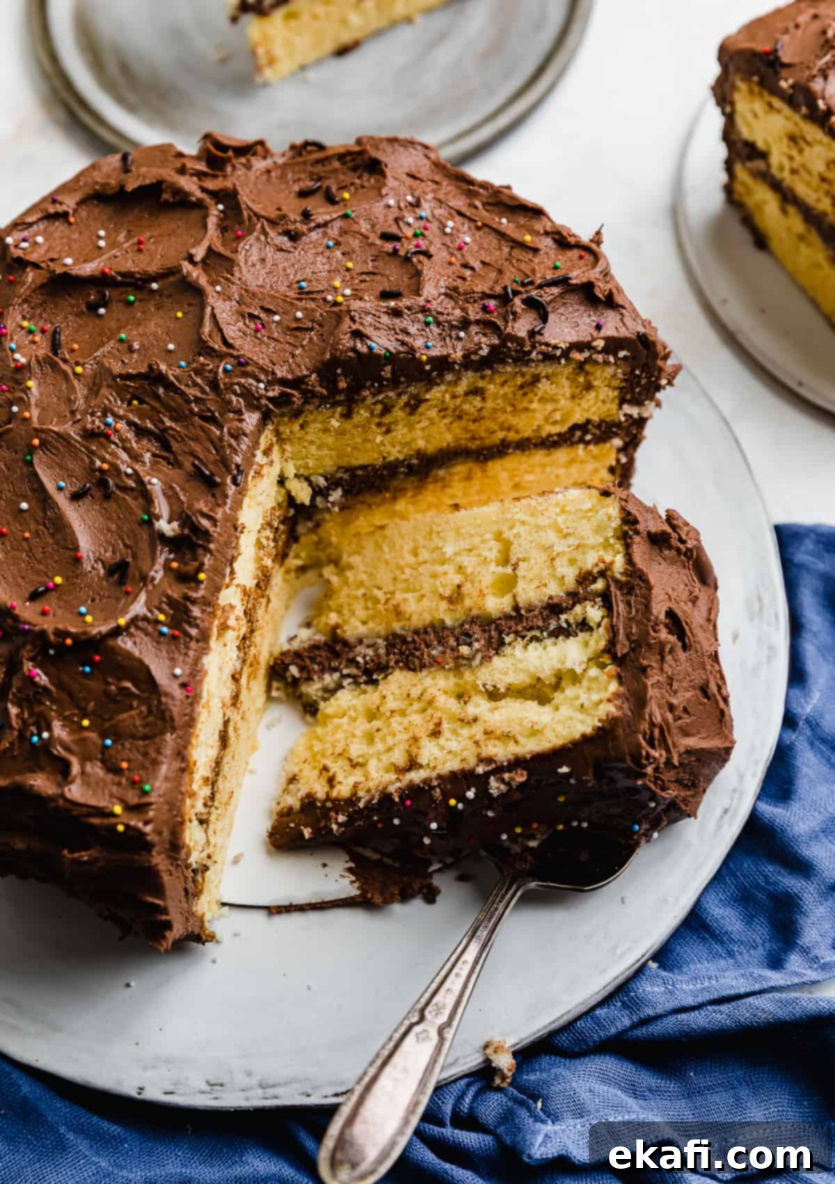 Golden Cake with Decadent Chocolate Frosting 2 Yellow layer cake with chocolate frosting on a white cake plate, with a slice of cake cut and laying flat on the platter.