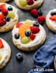 Fruity Dessert Cookie Pizzas 11 Fruit Pizza Cookies topped with a white cream cheese frosting and fresh fruit, on a black surface.
