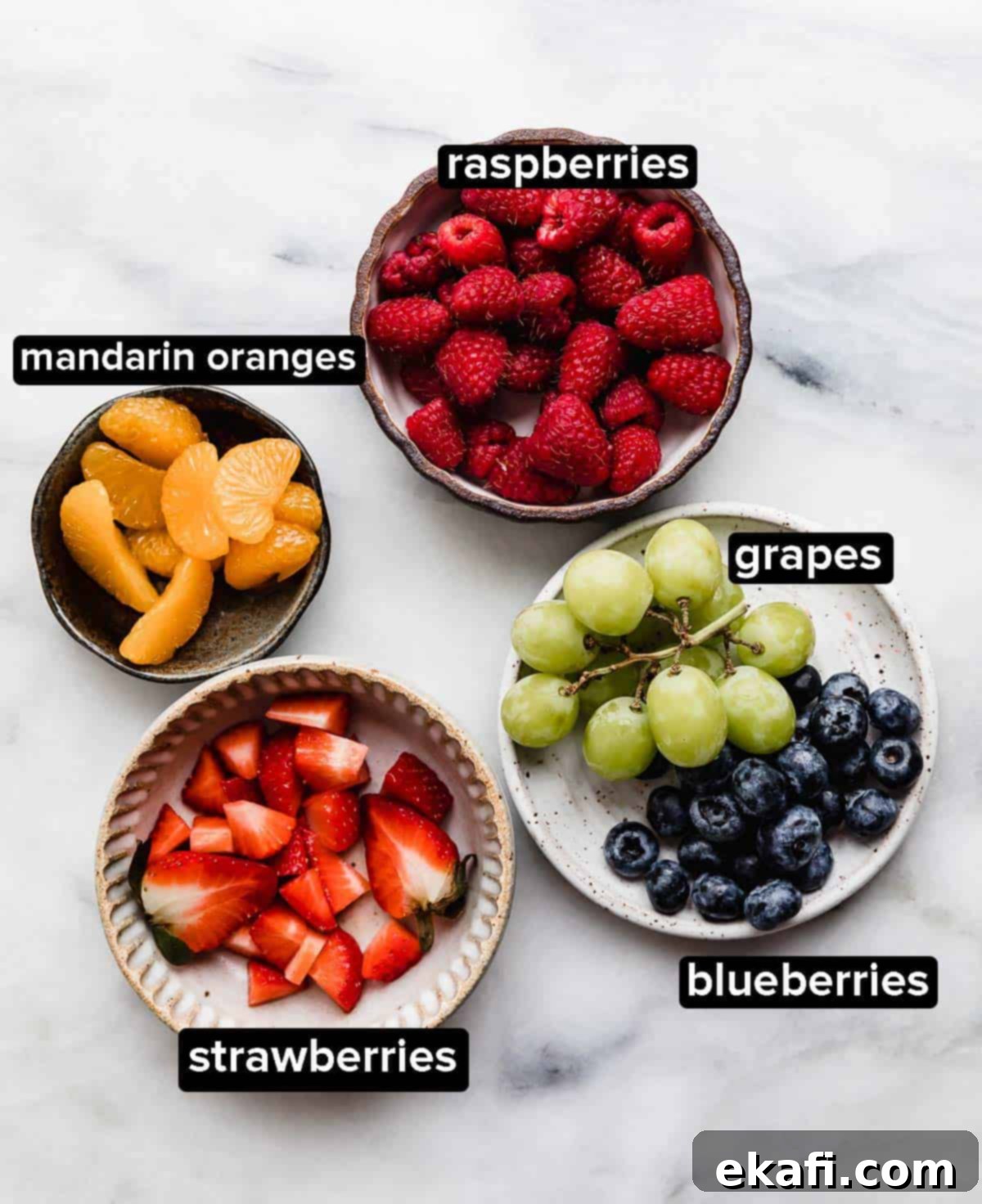 Fruity Dessert Cookie Pizzas 5 A vibrant selection of Fruit Pizza Cookies toppings: fresh raspberries, sweet mandarin oranges, sliced strawberries, juicy green grapes, and ripe blueberries, arranged on a white surface.
