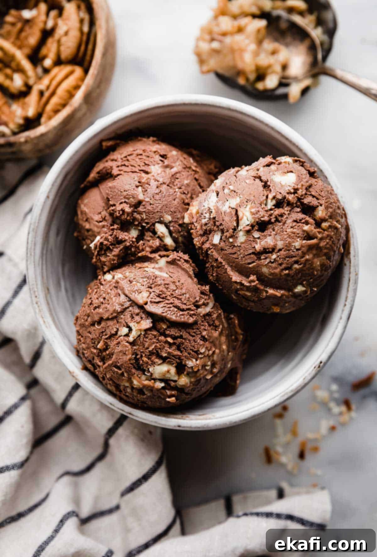 Decadent German Chocolate Ice Cream 2 Three scoops of dark chocolate German Chocolate ice cream in a white bowl with a white and blue stripe napkin to the left of the bowl.