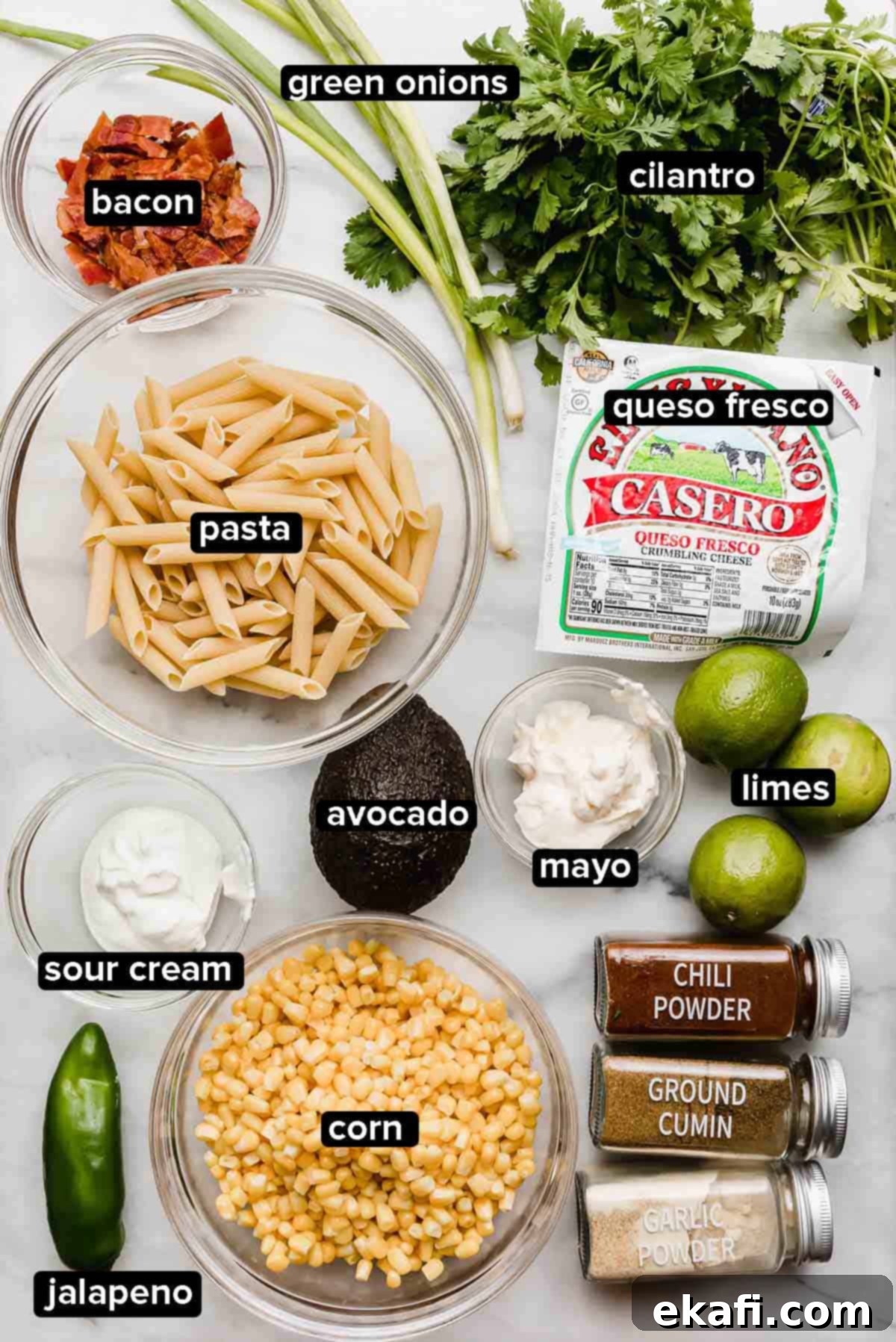 Elote Fiesta Pasta Salad 3 Mexican Street Corn Pasta Salad ingredients on a white marble surface: pasta, cilantro, jalapeño, corn, bacon, avocado, queso fresco, limes, and seasonings.