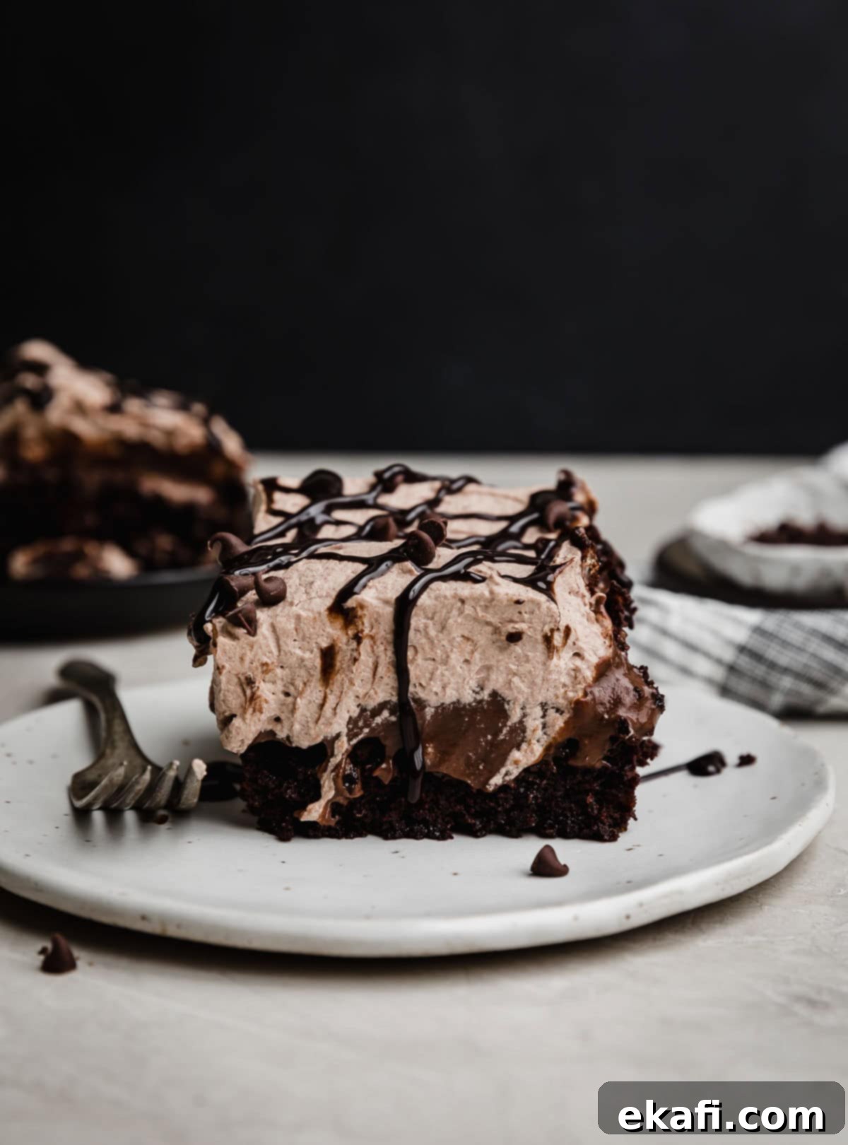 Chocolate Poke Cake 2 A slice of Triple Chocolate Poke Cake on a white plate against a dark background, showing layers of moist cake, rich pudding, and fluffy chocolate whipped cream, garnished with chocolate drizzle and mini chips.