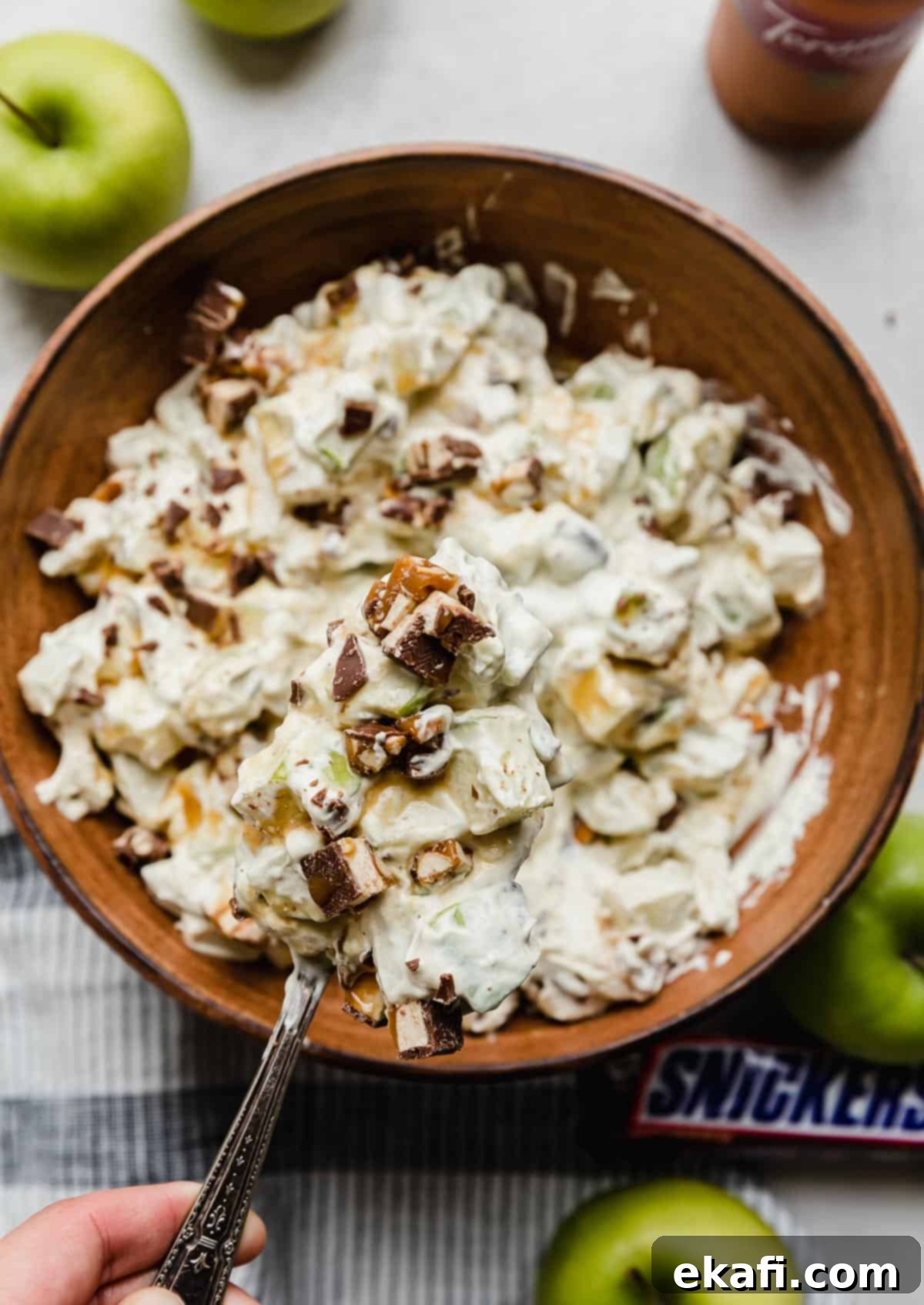 Creamy Snickers Apple Delight 2 A brown bowl filled with salad with apples and snickers and topped with caramel, on a white surface.