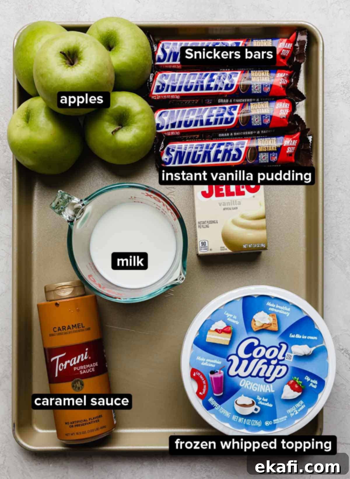 Creamy Snickers Apple Delight 3 Apple Snickers Salad ingredients on a baking sheet: snickers, cool whip, milk, vanilla pudding, apples, and caramel.