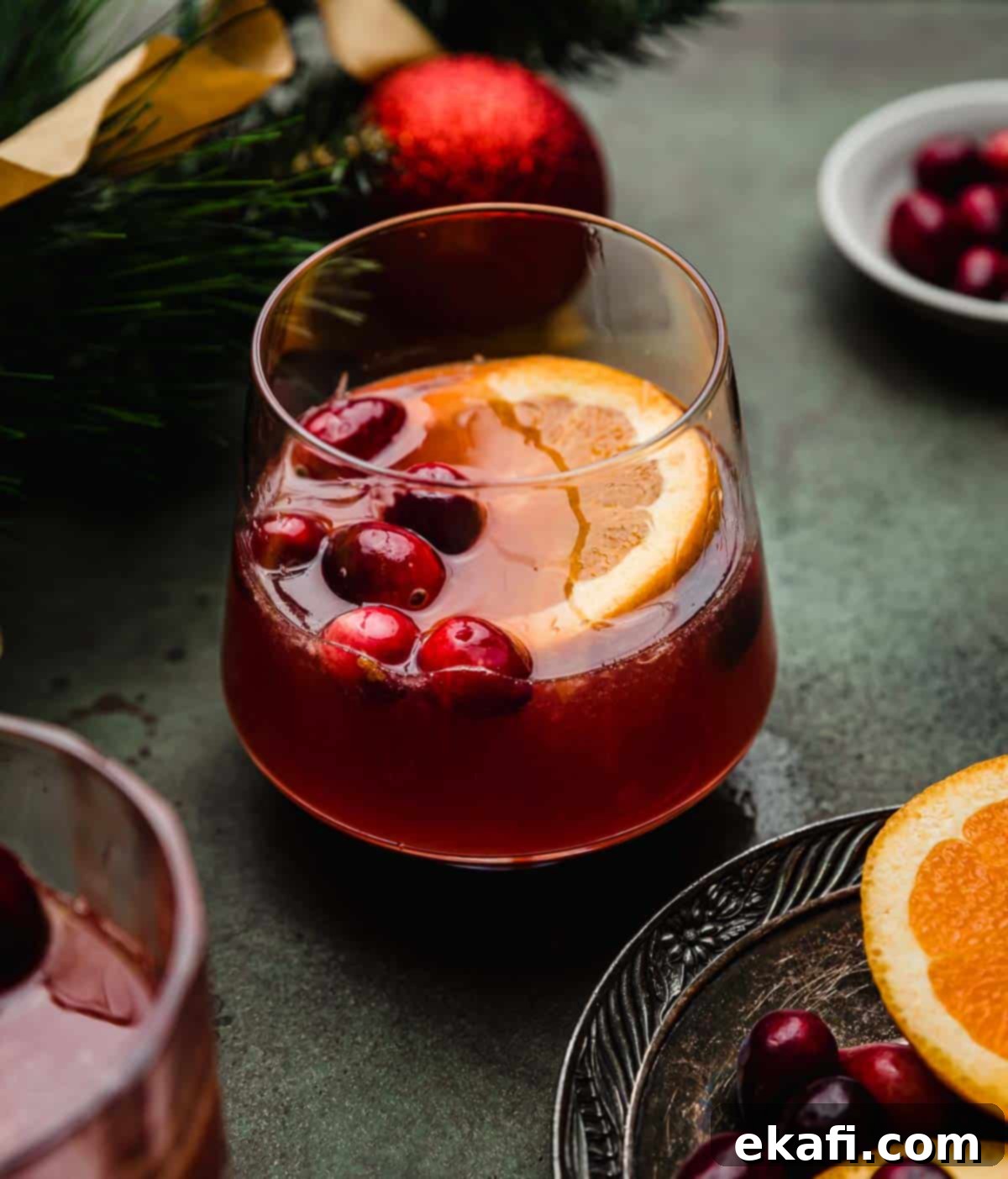Παρθένο Κράνμπερι Κοκτέιλ 2 Ένα Cranberry και πορτοκαλί Mocktail σε ένα γυάλινο ποτήρι με φρέσκα κράνμπερι και μια φέτα πορτοκαλιού, σε σκούρα πράσινη επιφάνεια.