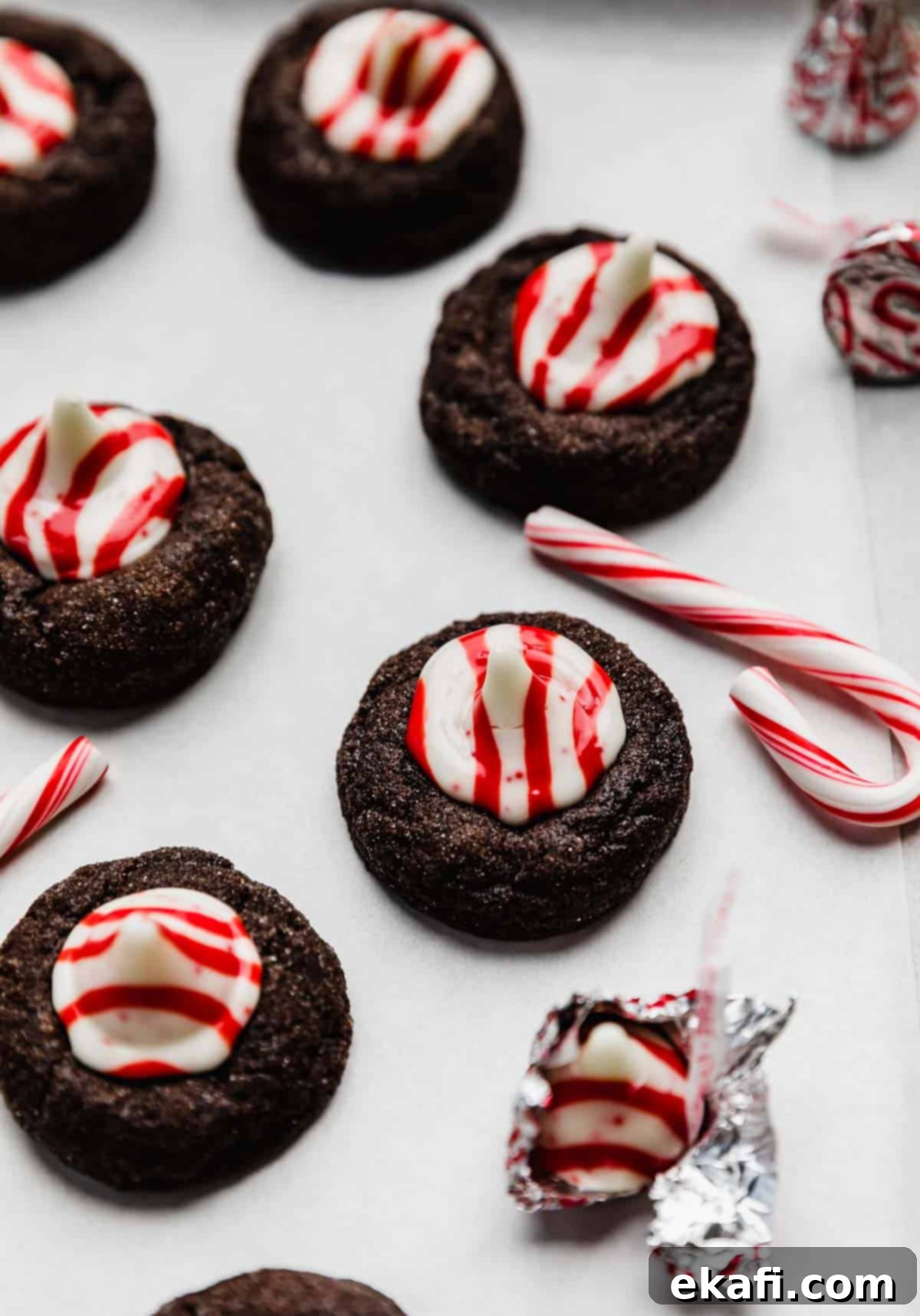 Peppermint Kissed Chocolate Gems 7 A chocolate cookie topped with a red and white striped peppermint candy kiss, on a white surface with a small candy cane next to the Christmas blossom cookie.