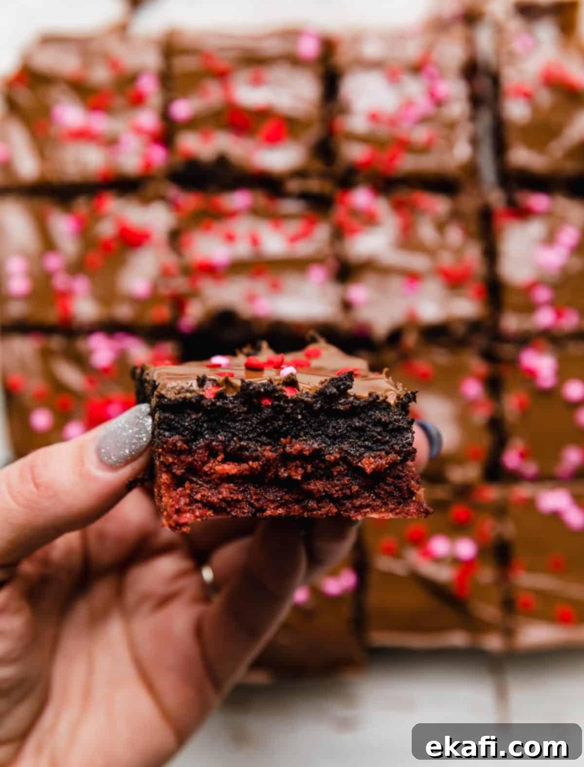 Crimson Oreo Truffle Dream Bars 8 A hand holding a Valentines brownie, featuring a red velvet base, Oreo truffle center, and melted milk chocolate overtop, showing the delicious layers.