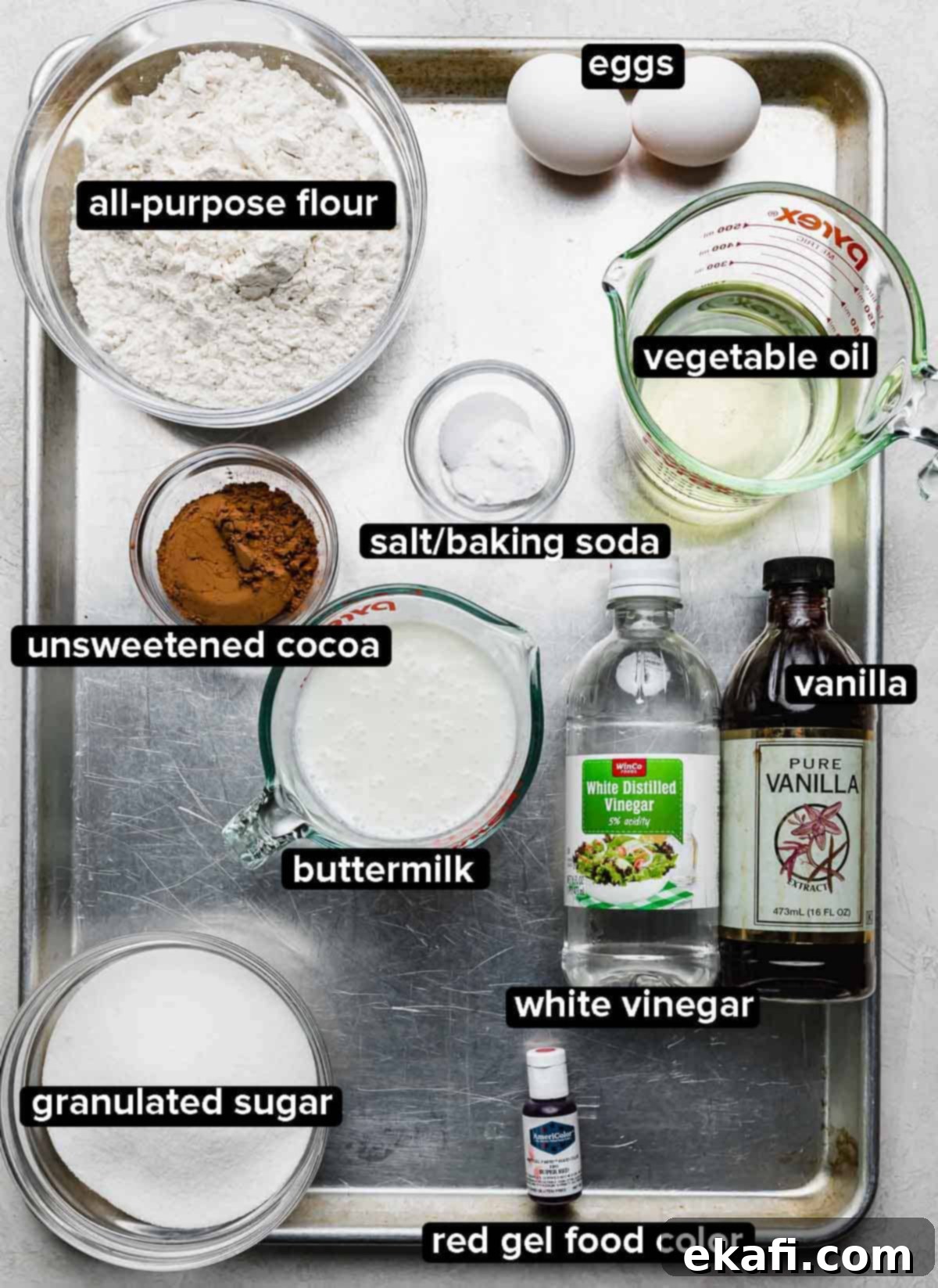 Velvety Crimson Crown 3 Red Velvet Bundt Cake ingredients portioned into glass bowls on a silver baking sheet: oil, buttermilk, vanilla, red gel food color, sugar, cocoa powder, eggs, salt and baking soda, and white vinegar, all ready for baking.