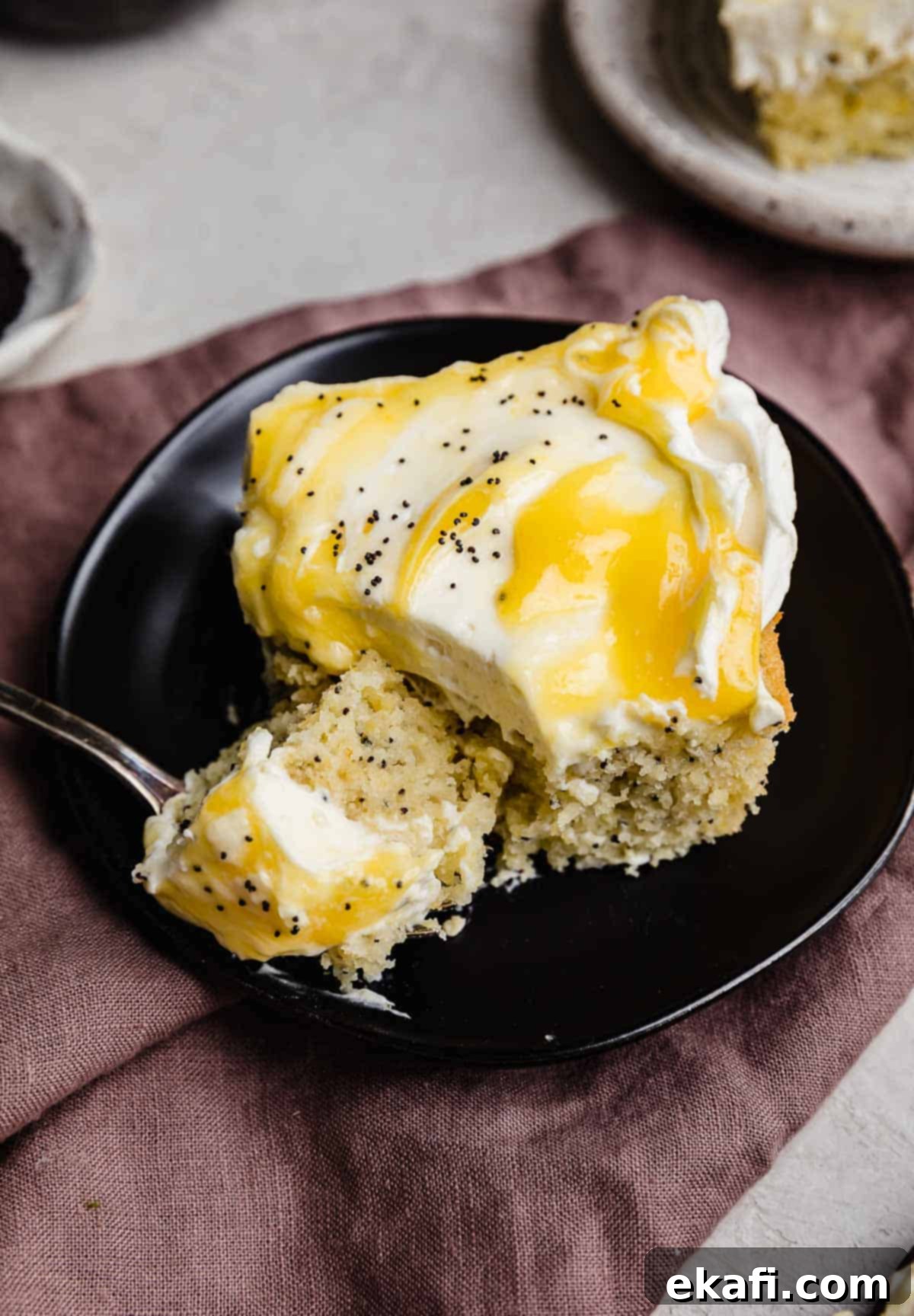 Sunshine Lemon Poppy Seed Cloud Cake 2 A square slice of Lemon and Poppy Seed Cake topped with whipped cream cheese frosting and lemon curd on a black plate.