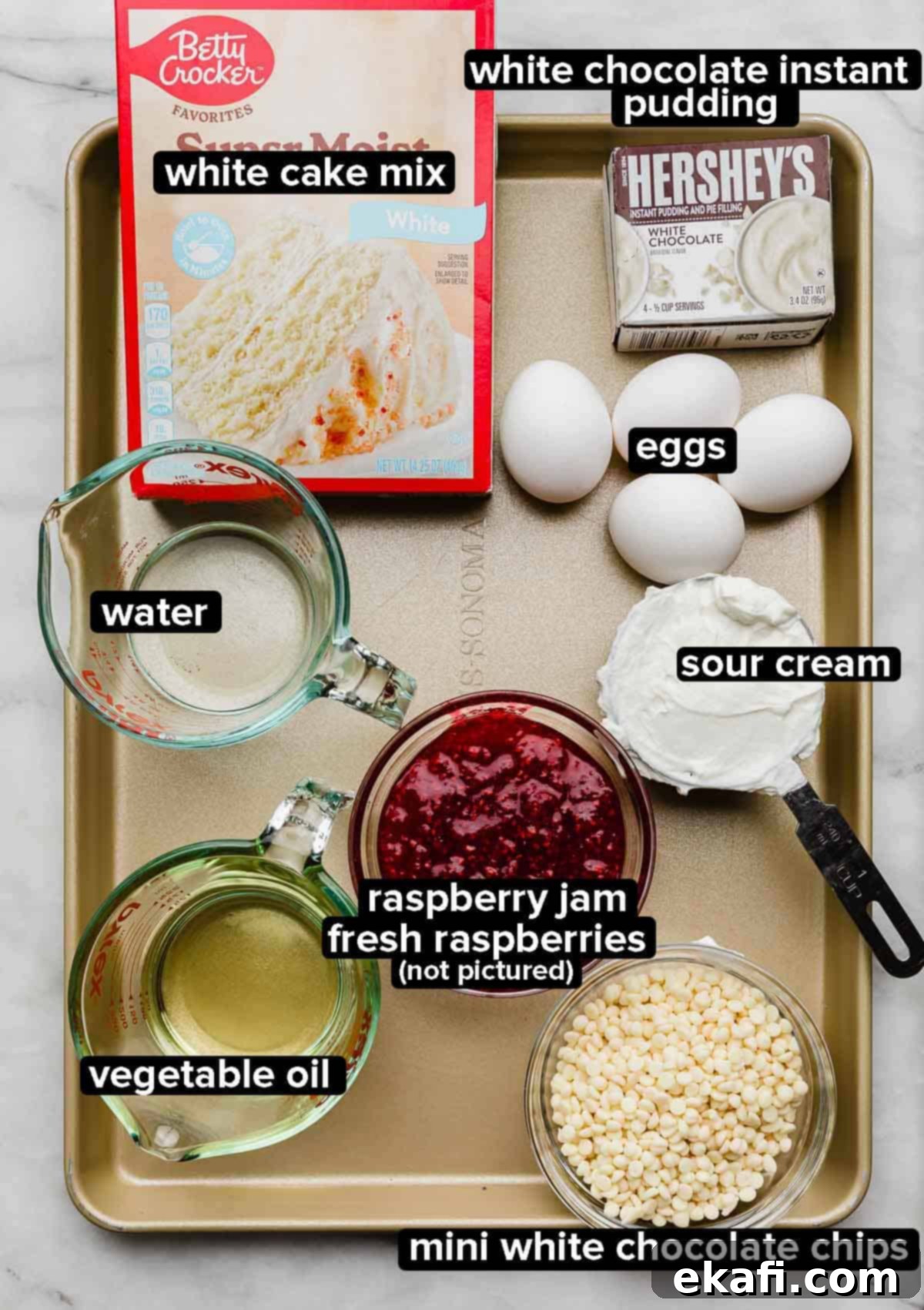 Decadent White Chocolate Raspberry Bundt Cake 3 White Chocolate Raspberry Bundt Cake recipe ingredients portioned into glass bowls on a bronze baking sheet: white cake mix, white chocolate pudding, white chocolate chips, raspberry jam, oil, water, eggs, and sour cream.