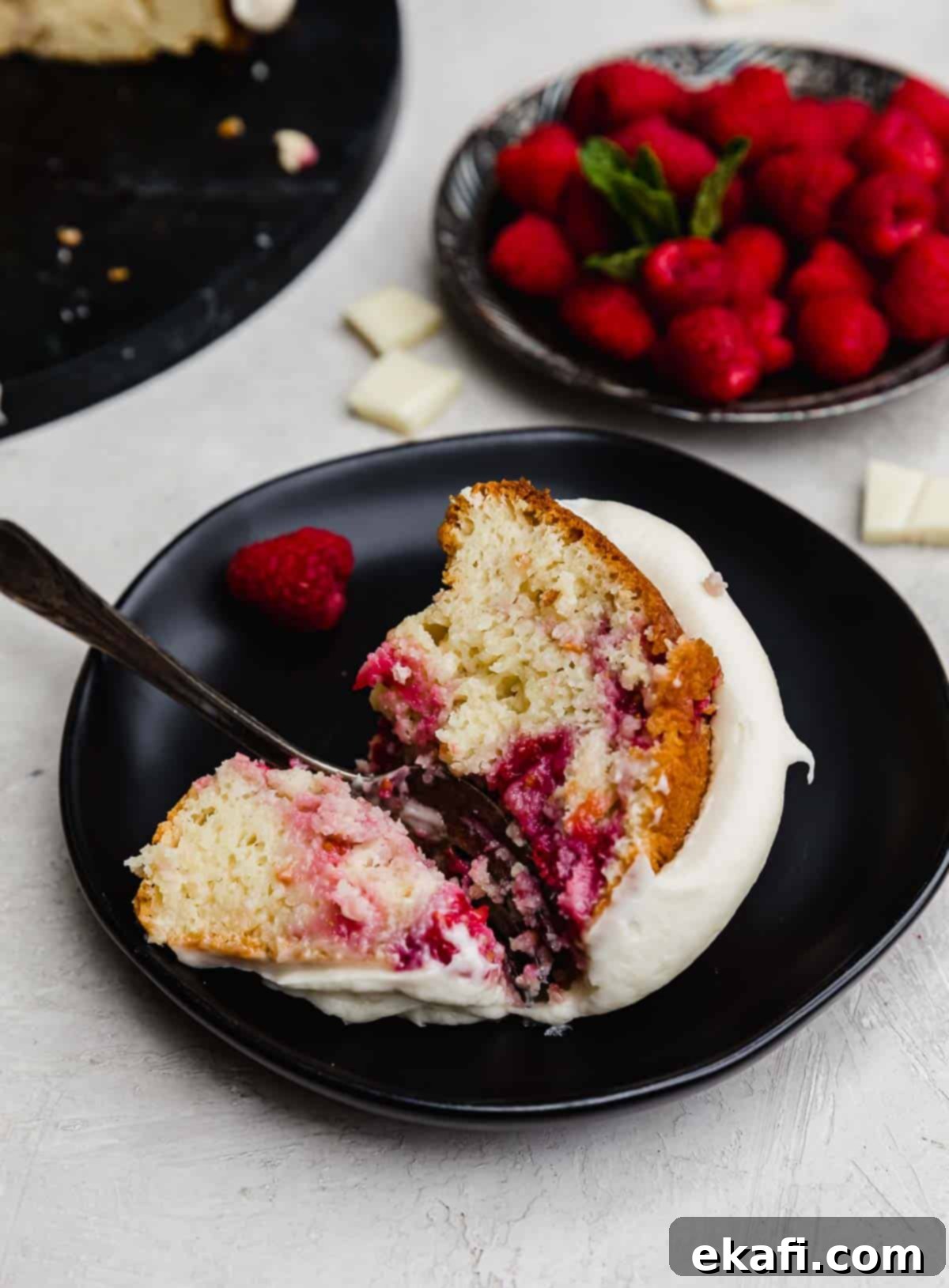 Decadent White Chocolate Raspberry Bundt Cake 7 A slice of White Chocolate Raspberry Bundt Cake topped with cream cheese frosting, with a slice on a black plate.