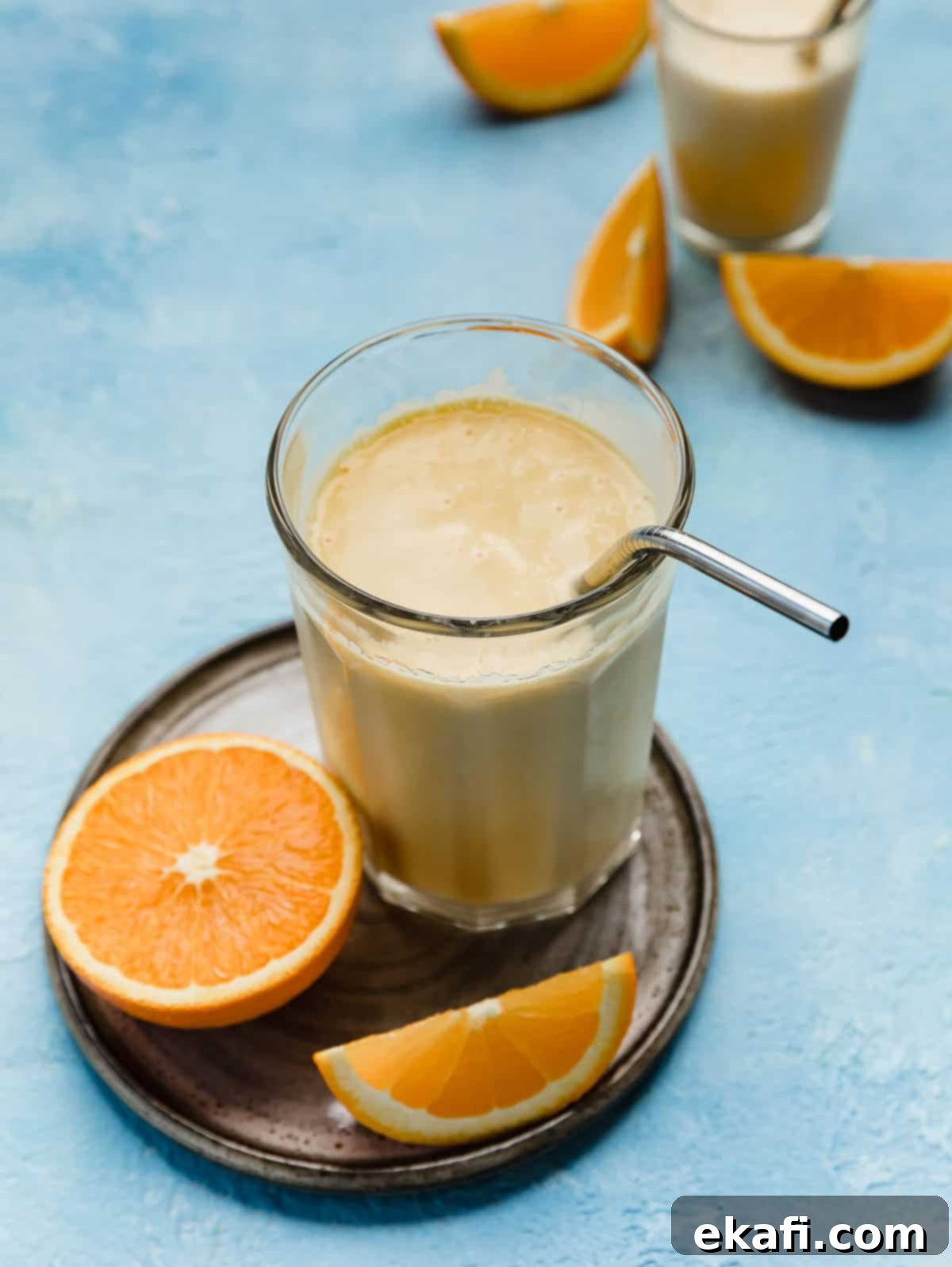 Βελούδινο Πορτοκάλι 2 Μια σπιτική συνταγή Orange Julius σε ένα μεγάλο γυάλινο ποτήρι με ένα μεταλλικό καλαμάκι, σε ανοιχτό μπλε φόντο.