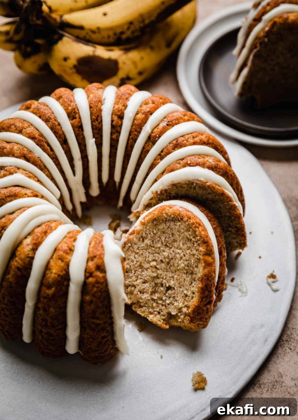 Classic Banana Bread Bundt Cake 2 A glazed Banana Bread Bundt Cake on a white cake plate with a slice of banana bundt cake tilting against the full cake.